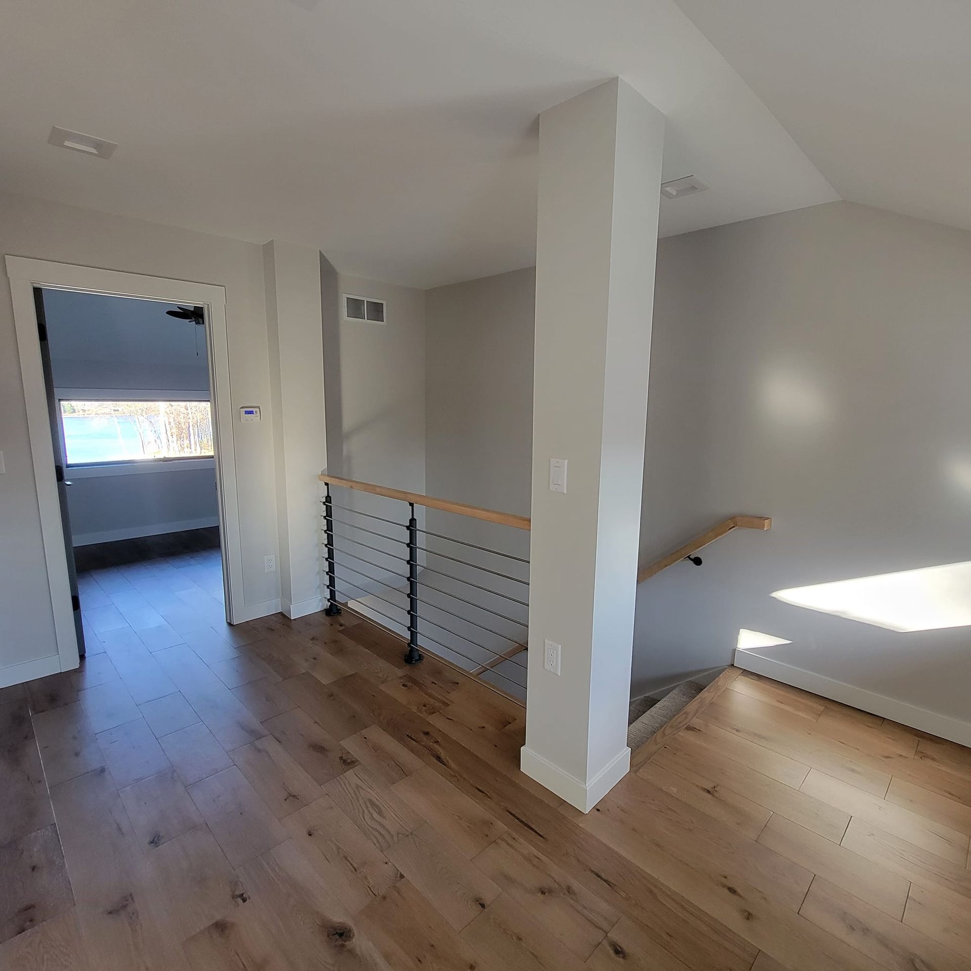 Interior view of a modern hallway with light wood flooring, a central structural column, and a cable railing staircase.