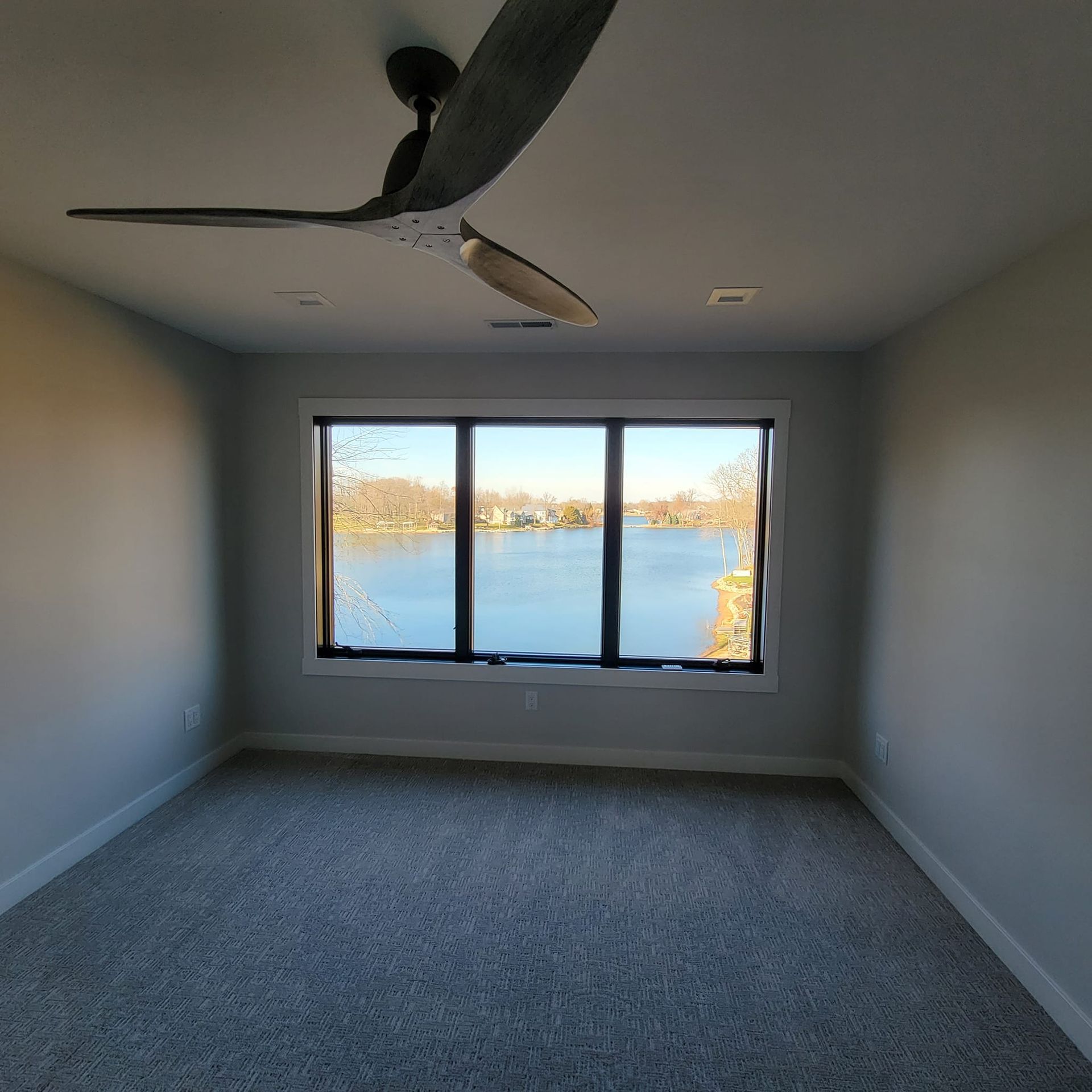 An empty, carpeted room with light gray walls, a modern ceiling fan, and a large window overlooking a calm lake.