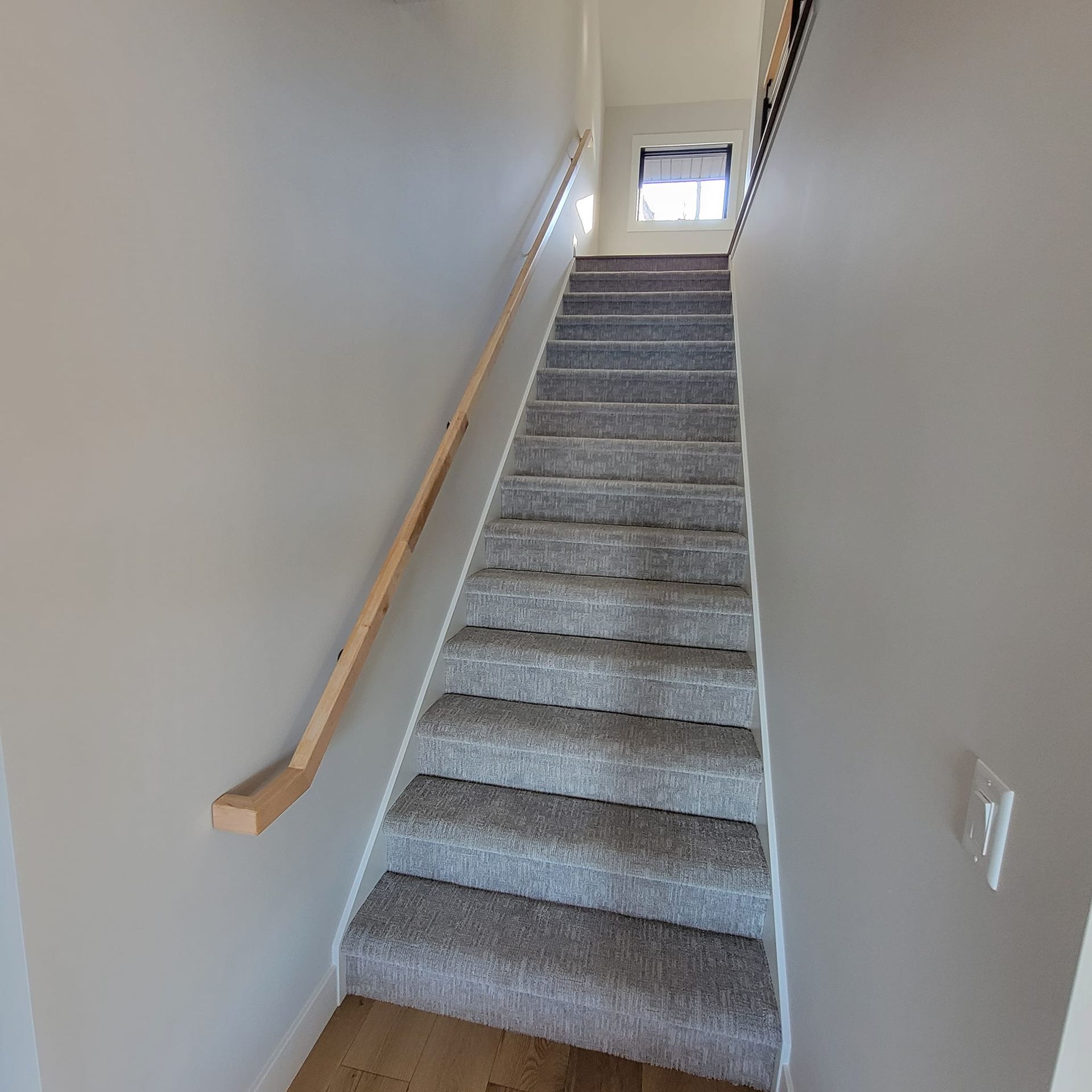 A set of straight carpeted stairs leads upward between two light gray walls, featuring a wooden handrail on the left.