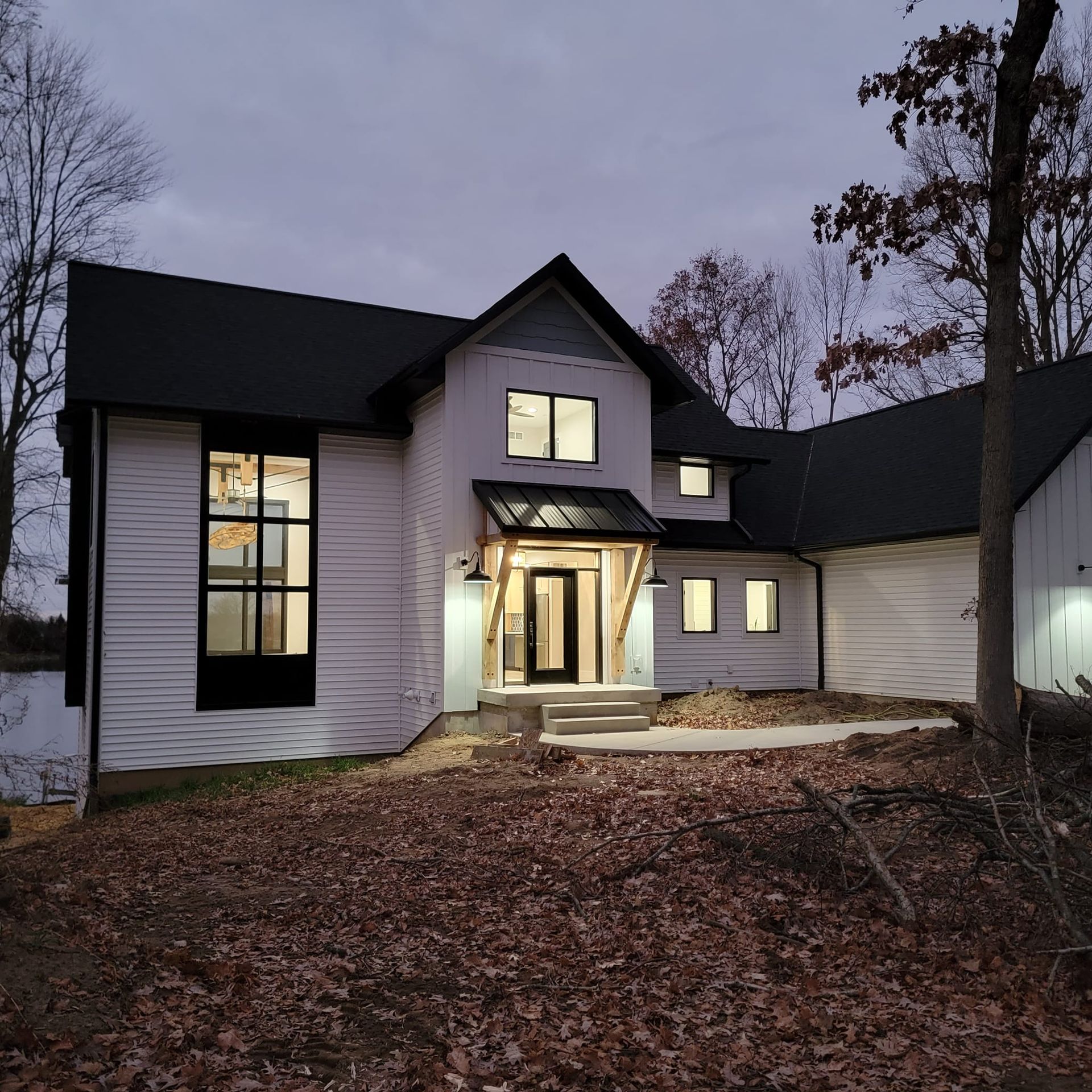 A white house with black trim and a wooden entryway porch at dusk, surrounded by trees and fallen leaves.