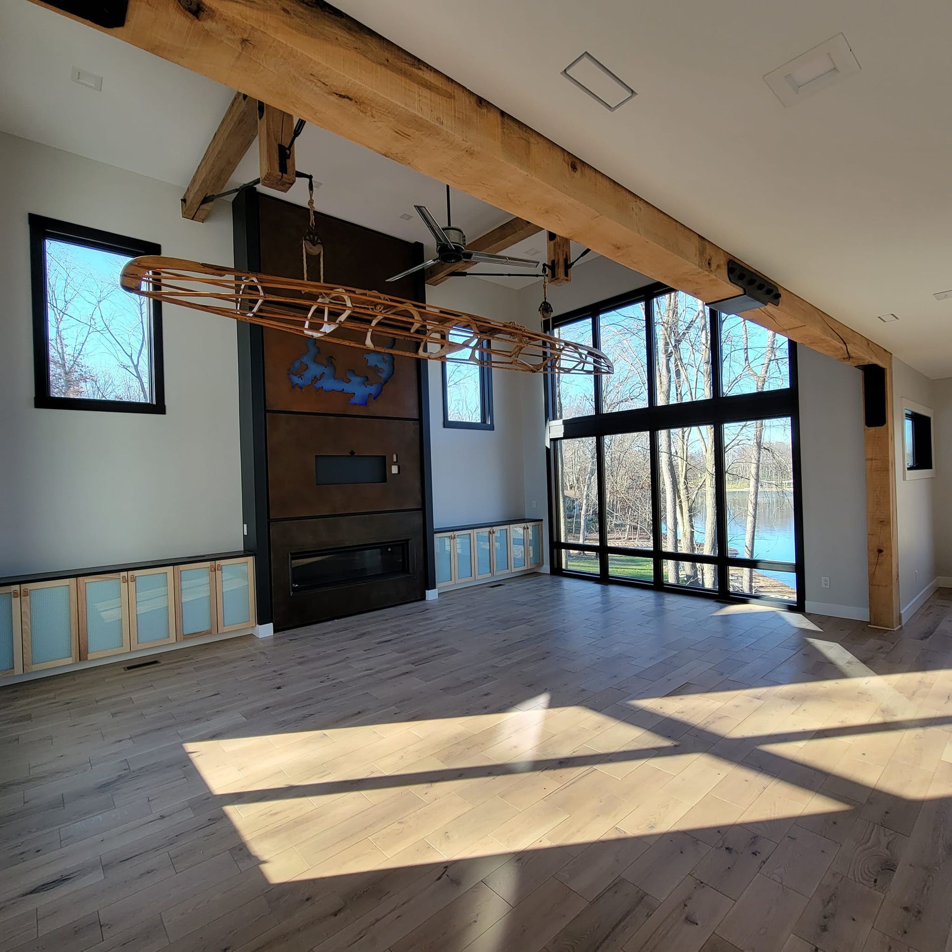 A spacious, modern room with light wood floors, a tall dark fireplace, large windows, and a hanging wooden light fixture.