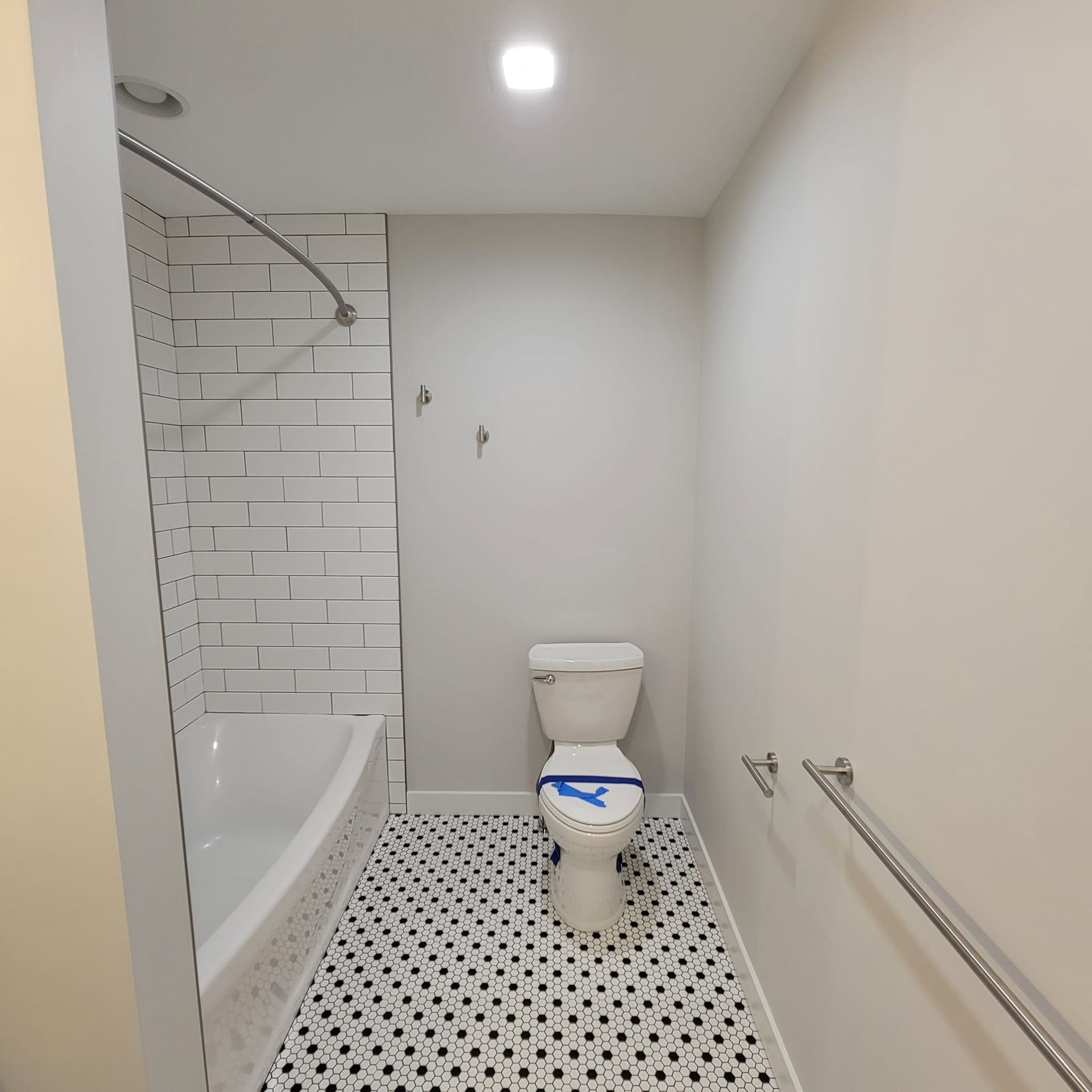 A small, modern bathroom with a white tiled tub, a toilet on a patterned black-and-white floor, and light gray walls.
