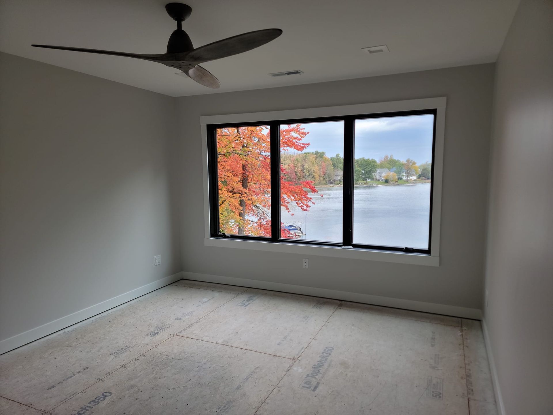 A room with gray walls, a black ceiling fan, and a large window overlooking a lake with vibrant autumn trees.