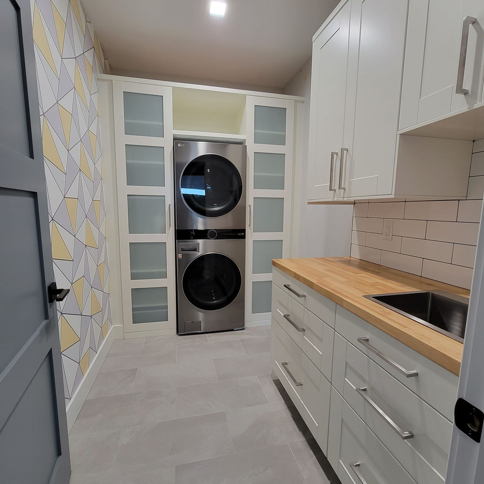 A modern laundry room with white cabinets, a wood countertop, a stacked washer and dryer, and patterned wallpaper.