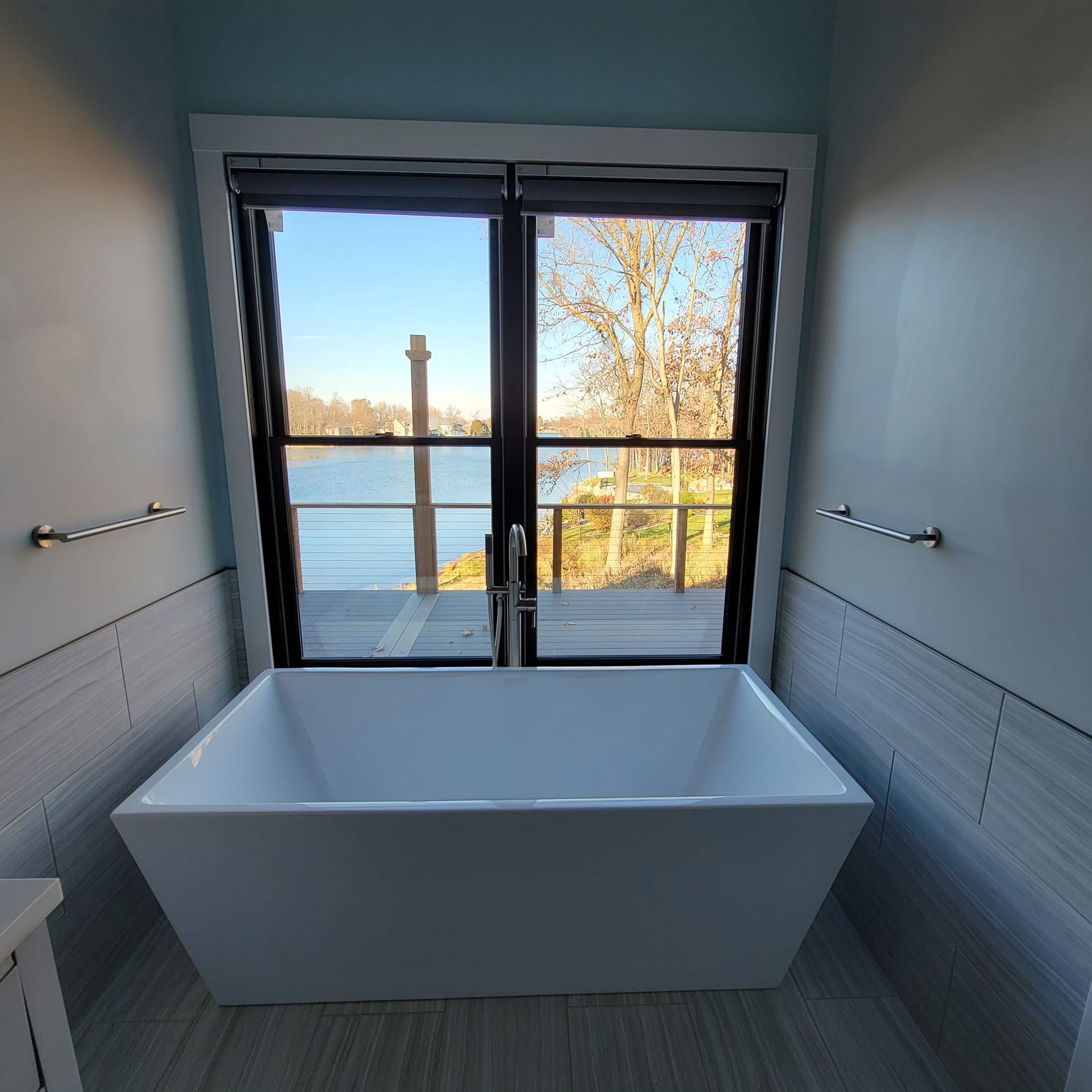 A white freestanding bathtub sits in front of a window overlooking a river, flanked by two silver towel bars on light walls.