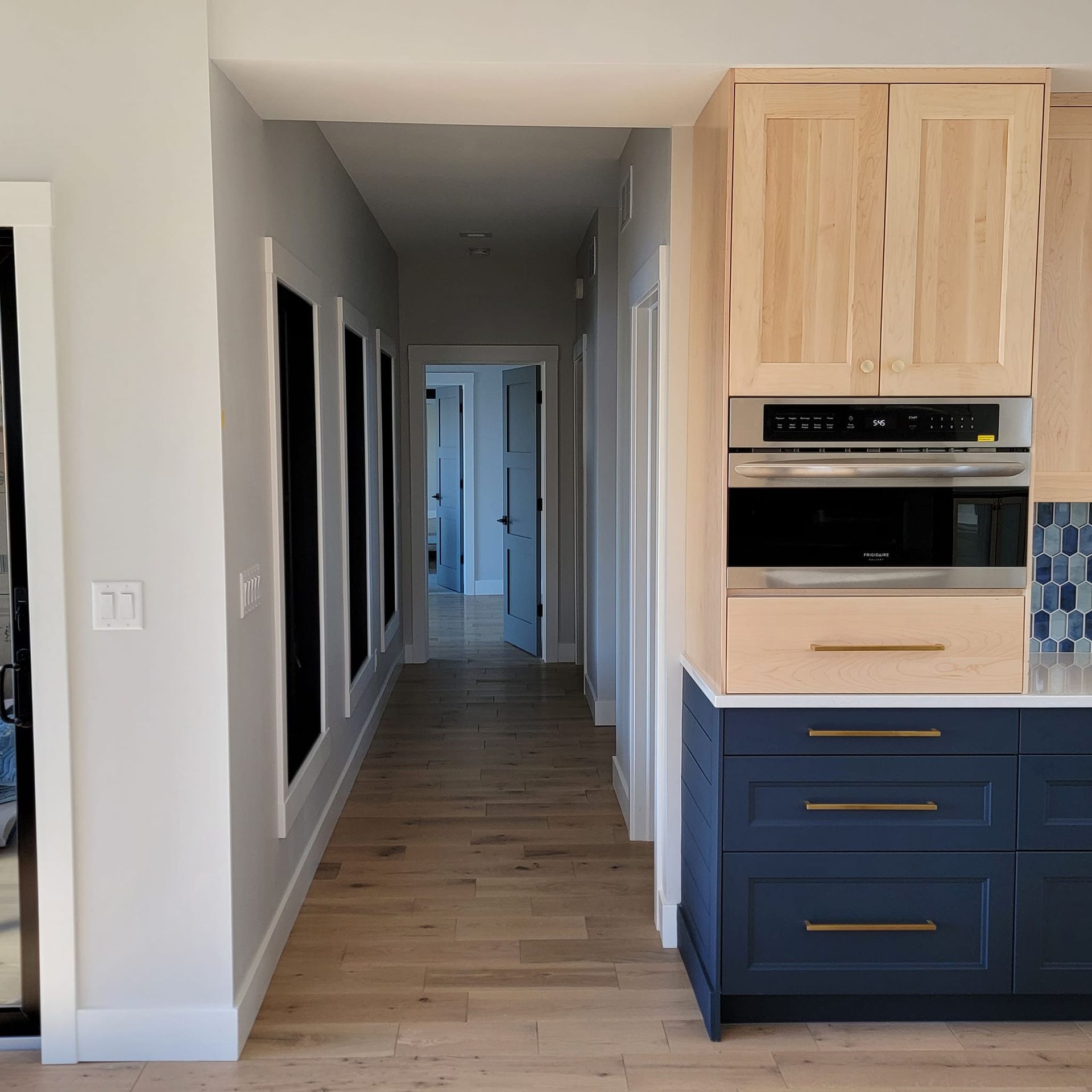 A hallway with light wood floors leading to an open door, next to a kitchen with navy lower and light wood upper cabinets.