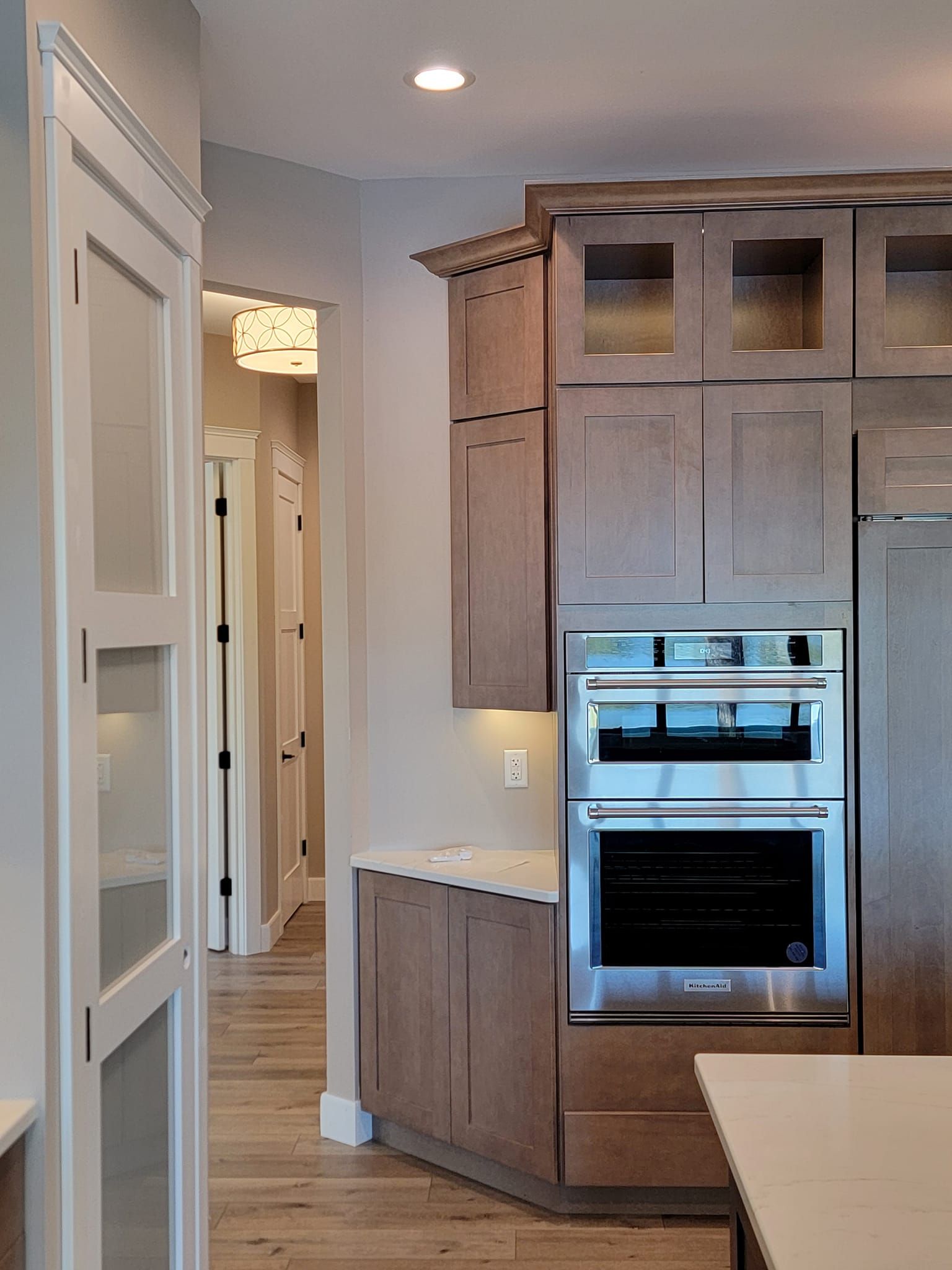 A modern kitchen with brown cabinets, a built-in wall oven, and a view into an adjacent hallway with light wood floors.