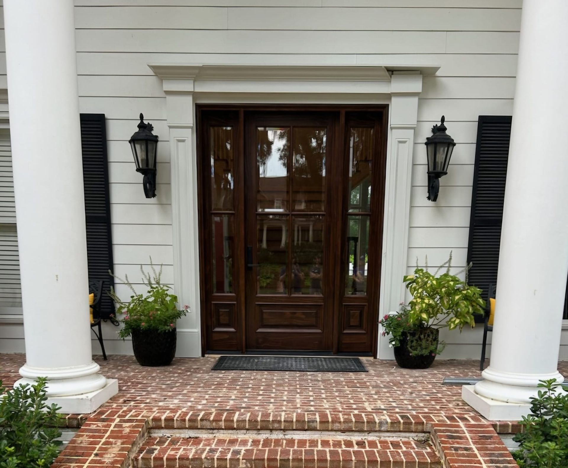 A front entrance with a dark wood door, sidelights, and black lanterns on white siding, framed by columns above brick steps.