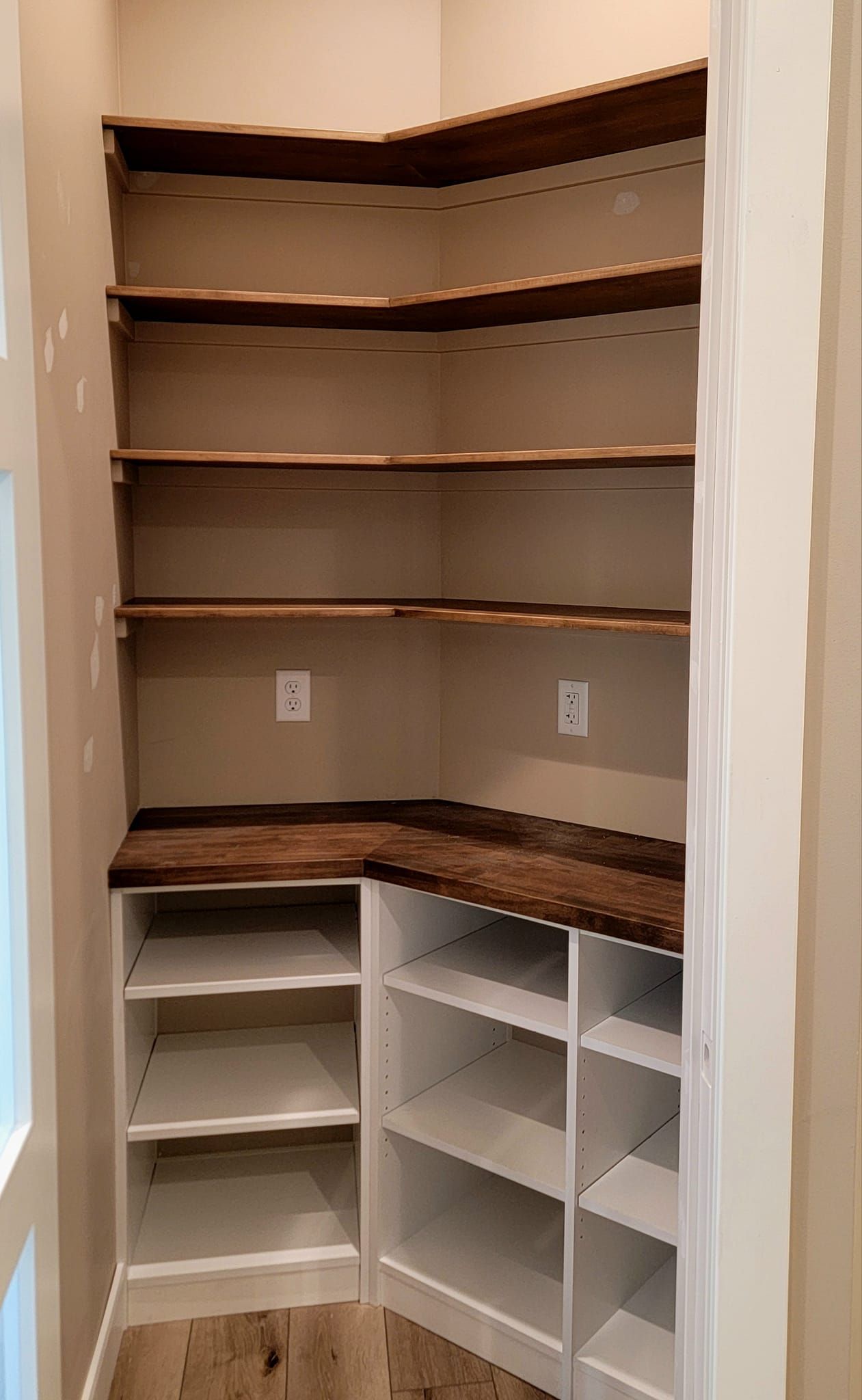 A corner walk-in pantry featuring white lower shelving units, a dark wood countertop, and upper floating wood shelves.