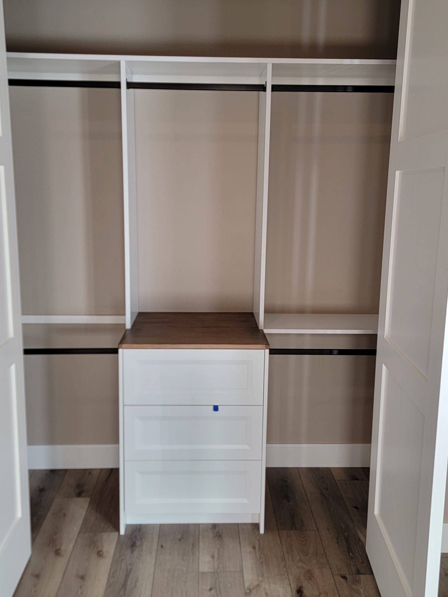A white reach-in closet with three shelves, hanging rods, and a central white three-drawer dresser with a wooden top.