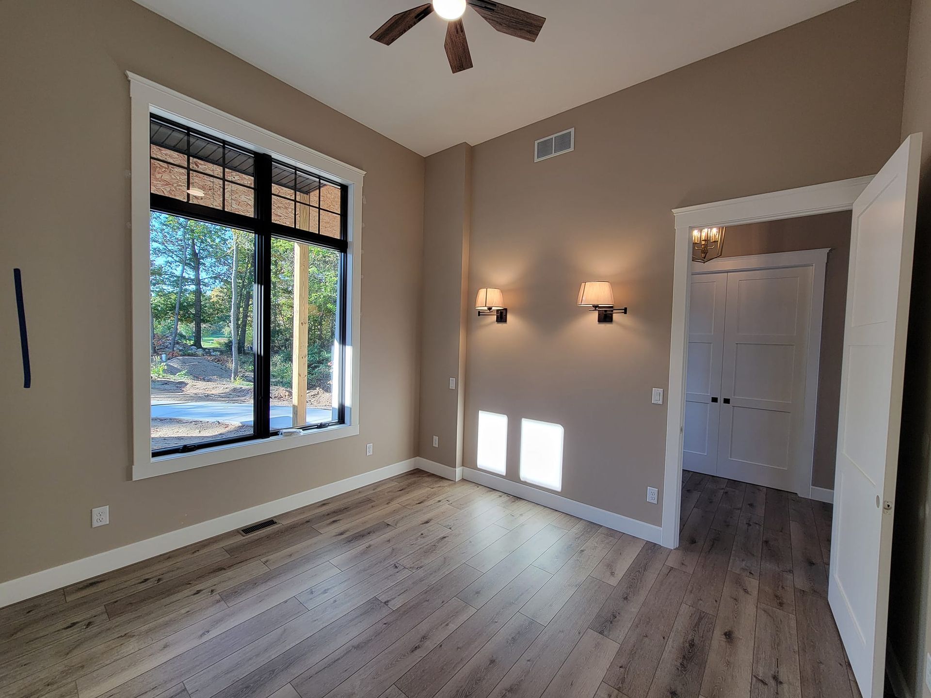 A bright, empty room with light wood floors, tan walls, a large window, two wall sconces, and an open doorway.