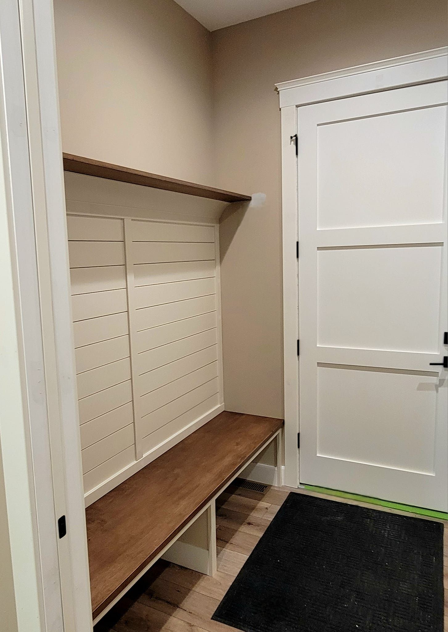 A white slatted mudroom bench with a wood seat and upper shelf next to a white door in a neutral-toned entryway.