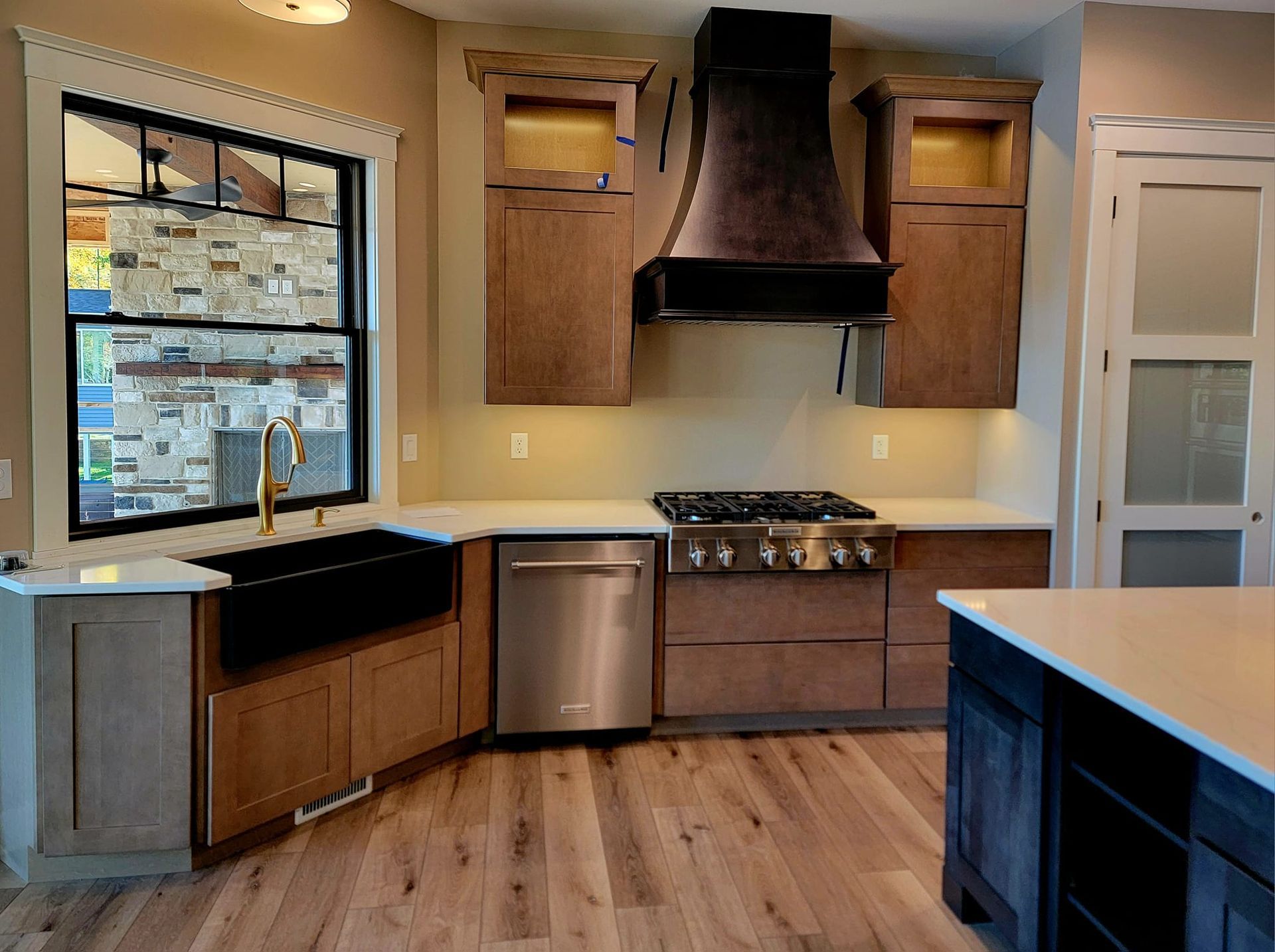 A modern kitchen with wood cabinets, white countertops, a black farmhouse sink, and a stainless steel stove with a hood.