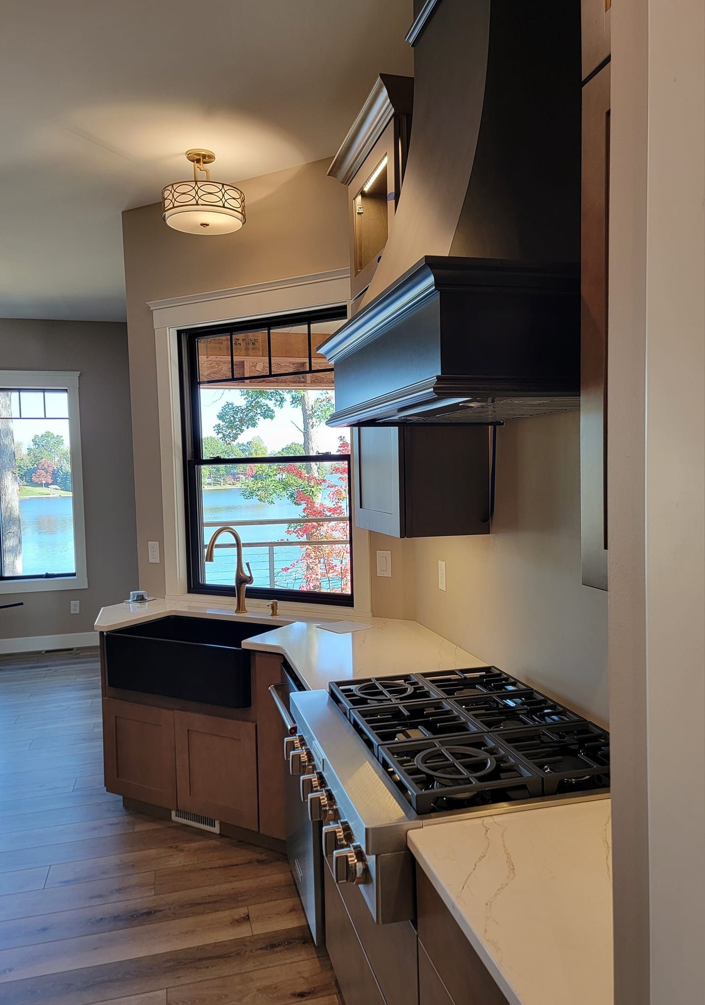 A modern kitchen with white countertops, a black farmhouse sink, a stainless gas range, and a view of a lake.