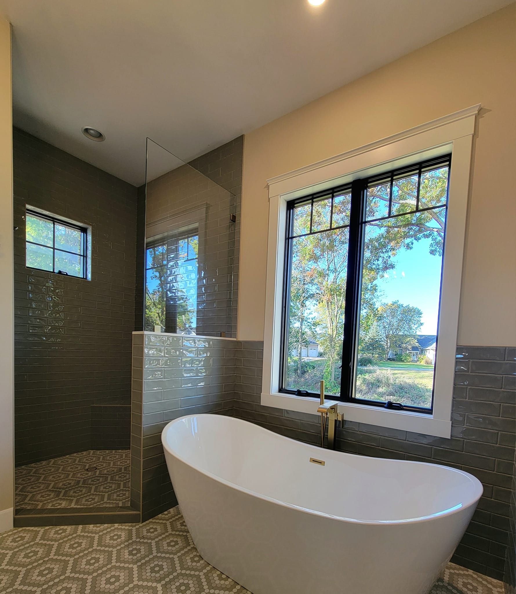 A modern bathroom featuring a white freestanding tub, a large window, and a walk-in shower with grey patterned tile.