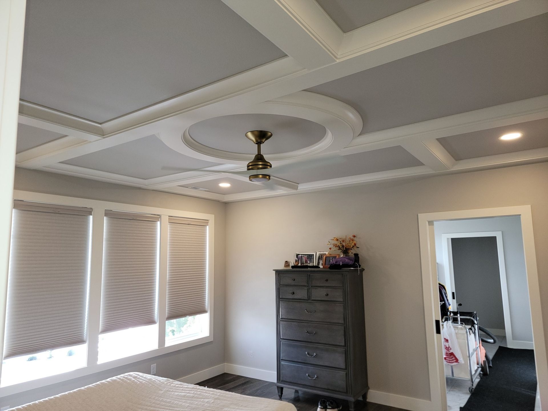 A bedroom with light gray walls, a coffered ceiling with a circular molding and fan, and windows with neutral shades.