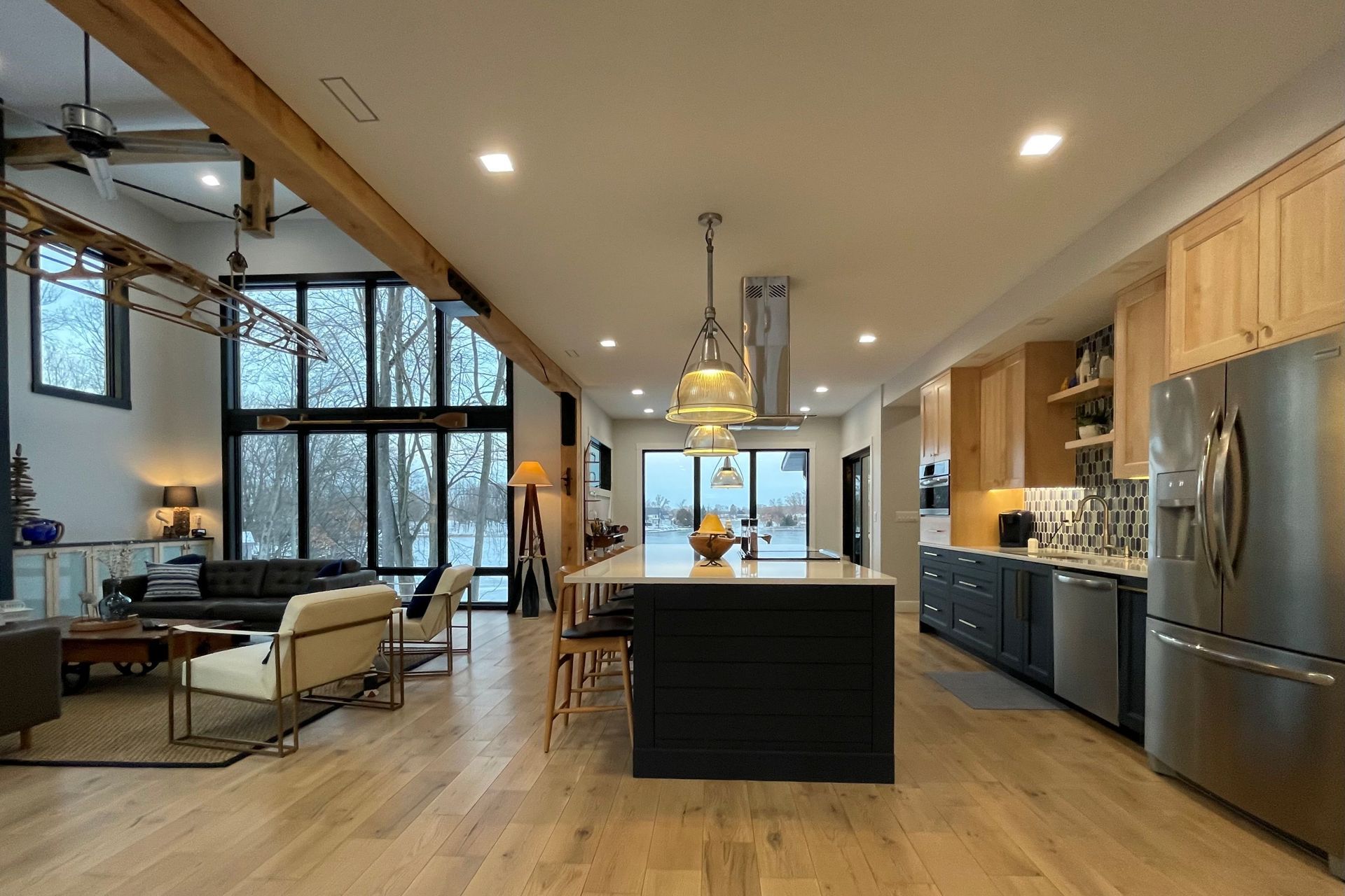 A modern, open-concept kitchen and living area featuring a large island, stainless steel fridge, and tall glass windows.