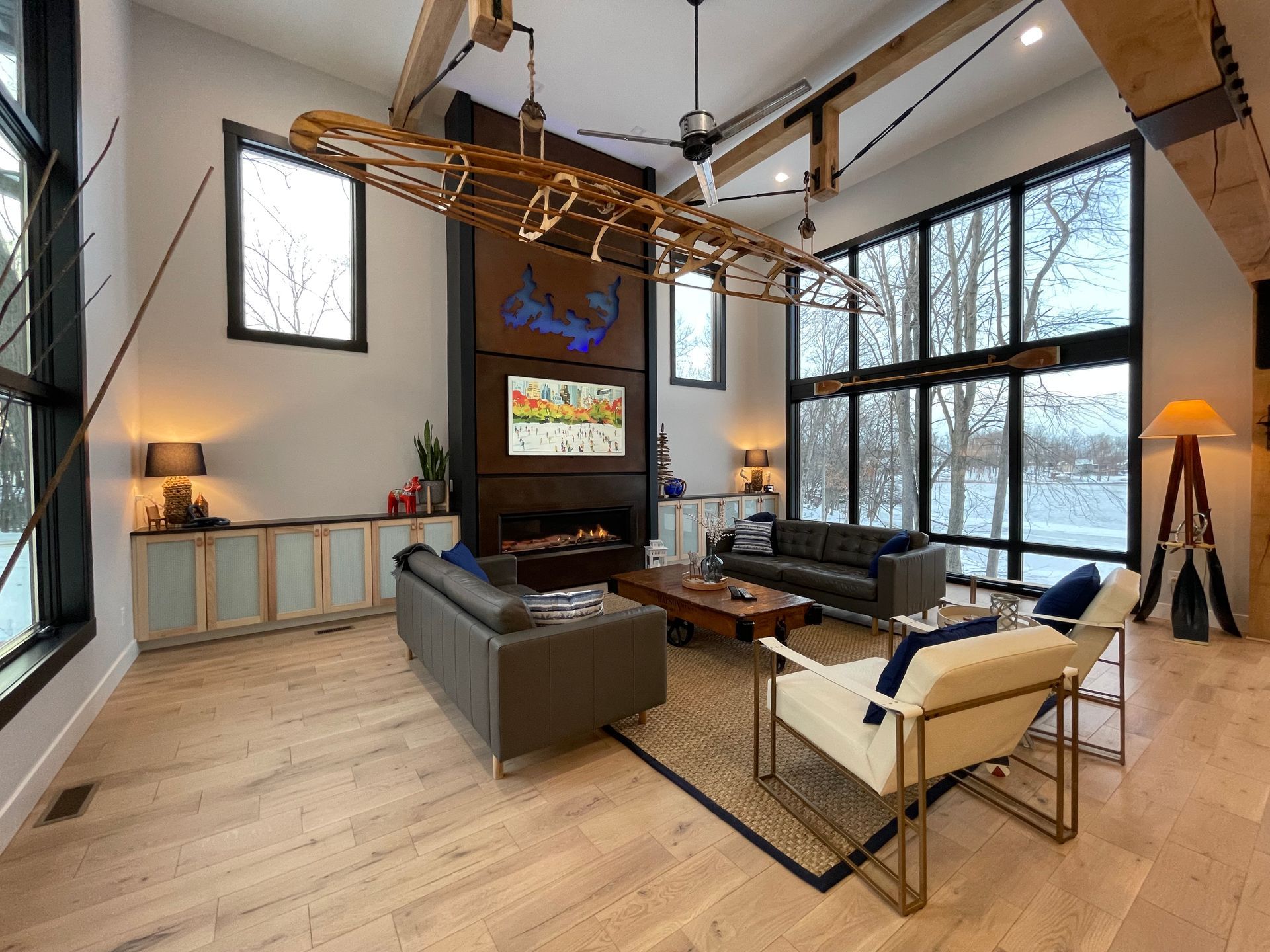 Modern living room with a suspended canoe, fireplace, leather furniture, and large windows overlooking a snowy landscape.