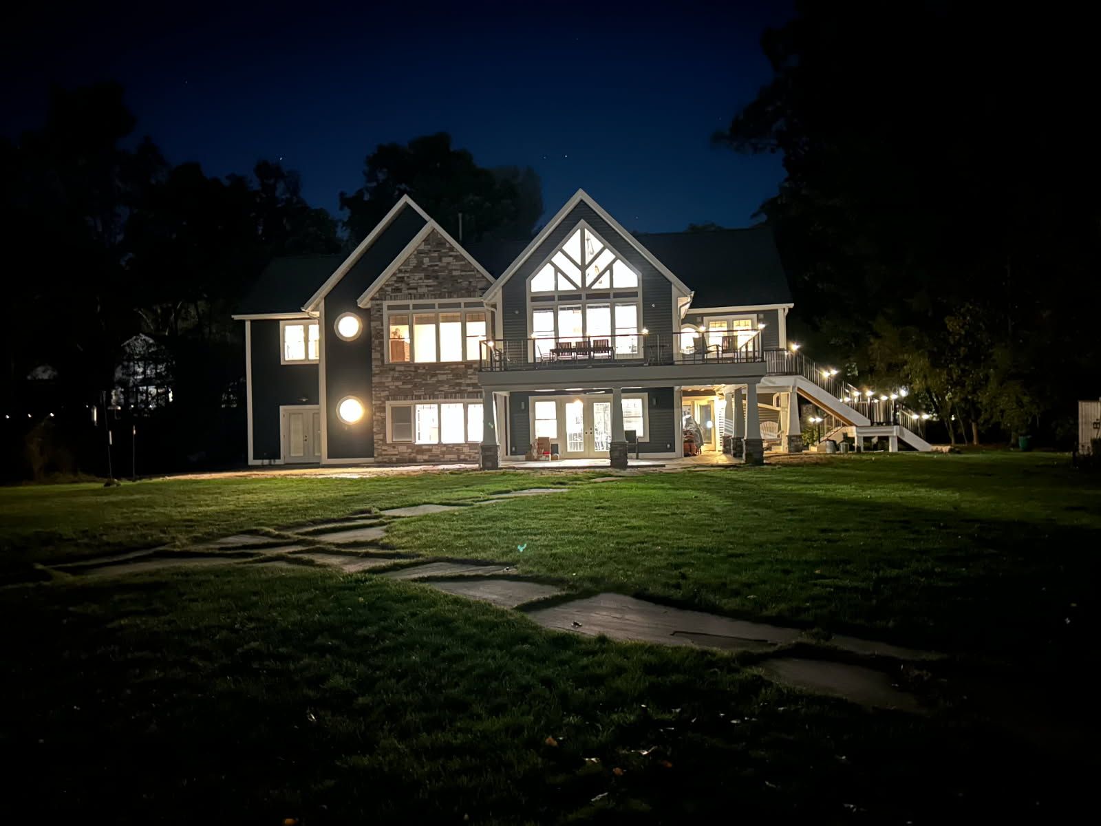 A multi-story house with illuminated windows and a stone exterior at night, set against a dark sky and a grassy lawn.