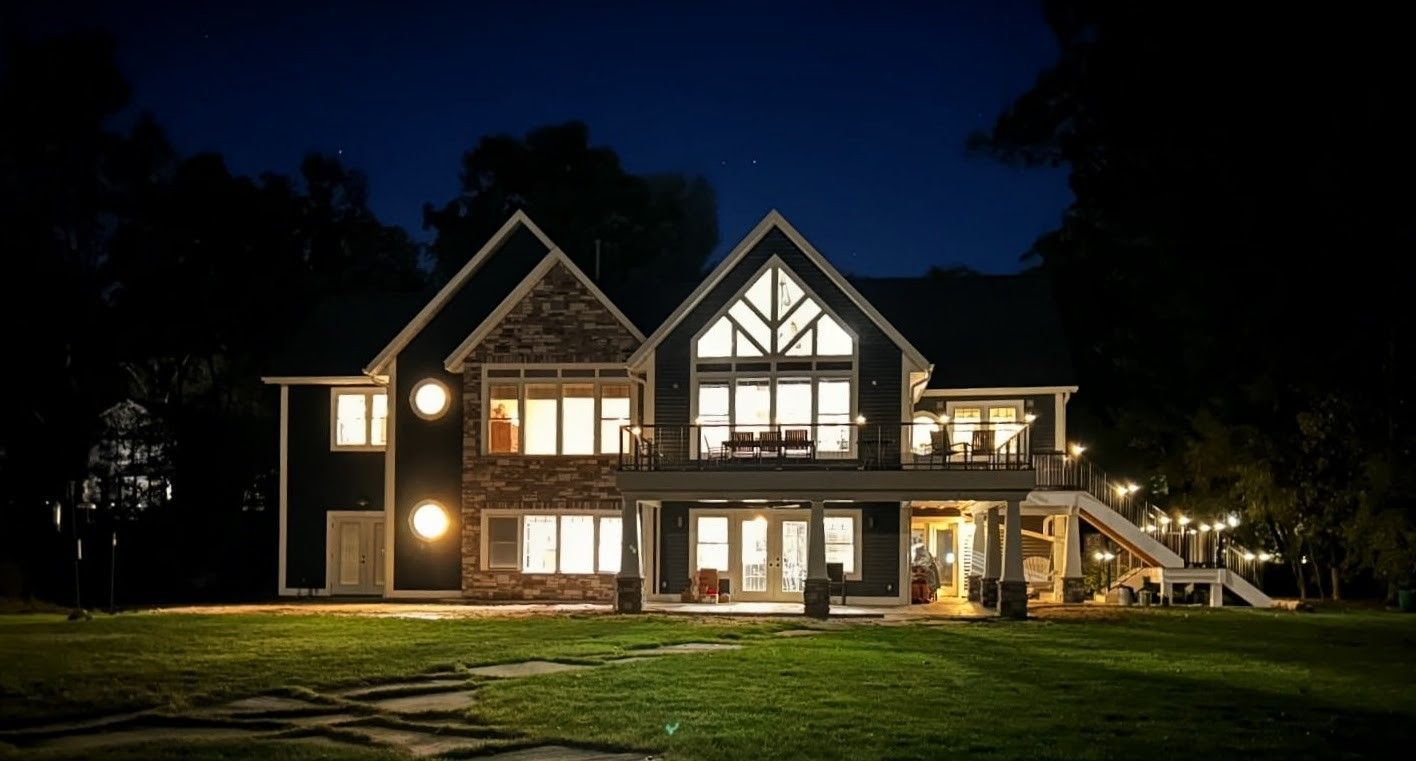 A two-story dark-colored house with stone accents, illuminated by bright interior lights at night against a dark sky.