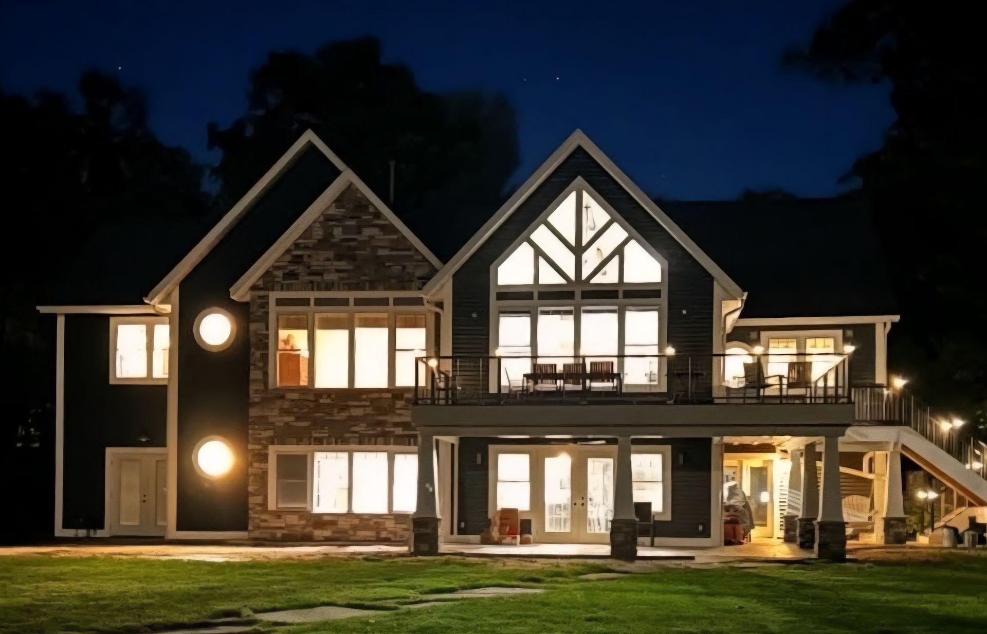A two-story dark-sided house with stone accents and illuminated windows glows at night against a dark sky.