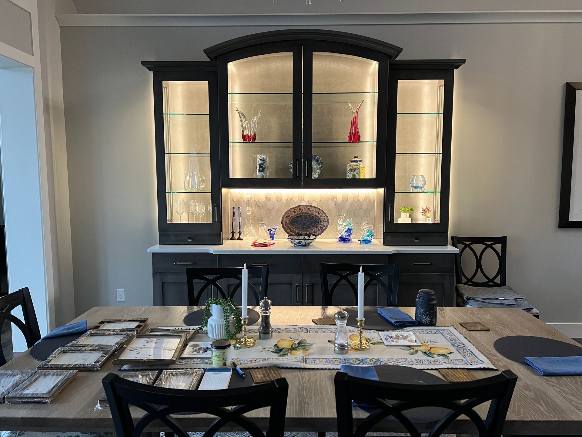 A dining room featuring a wooden table set for dinner in front of a dark, built-in display cabinet with interior lighting.