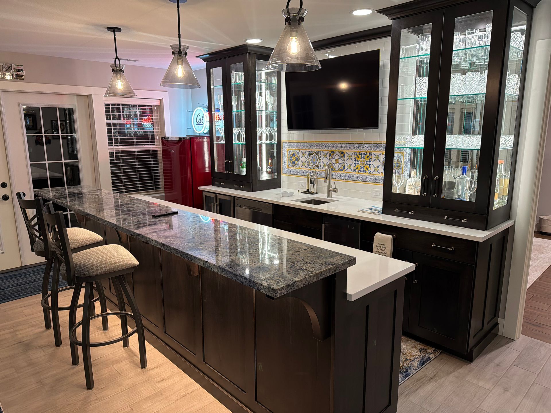 A home bar with granite counters, dark wood cabinets, glass display cases, bar stools, and a television on the wall.
