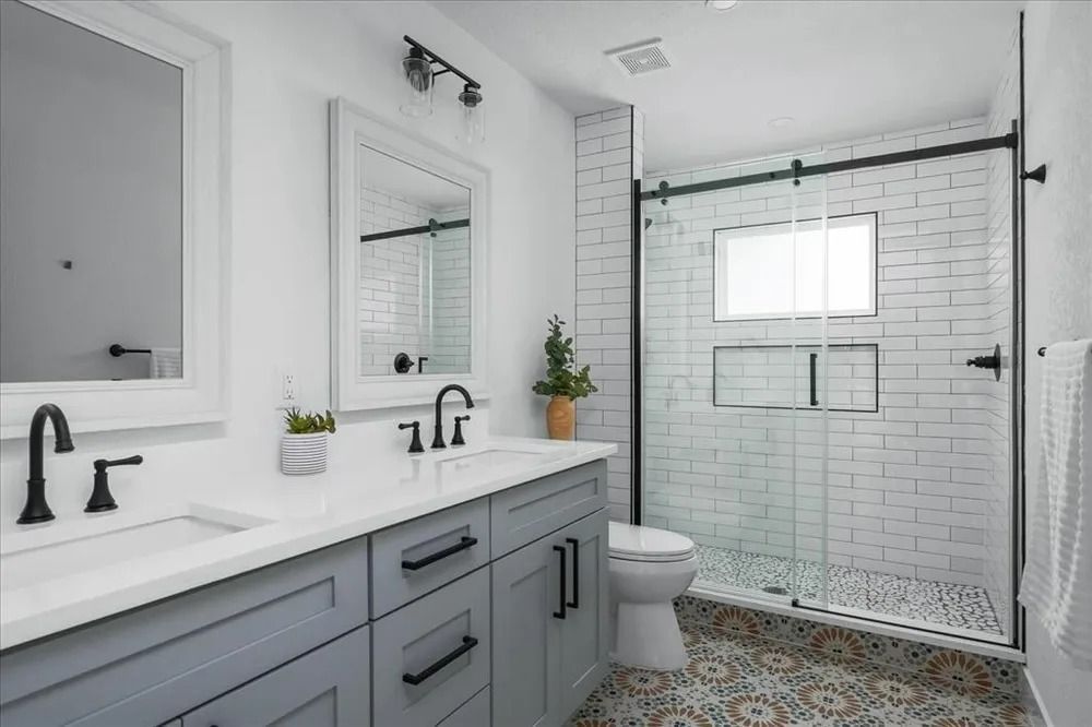Modern bathroom with a double vanity featuring gray cabinets, white countertops, black fixtures, and a glass shower.