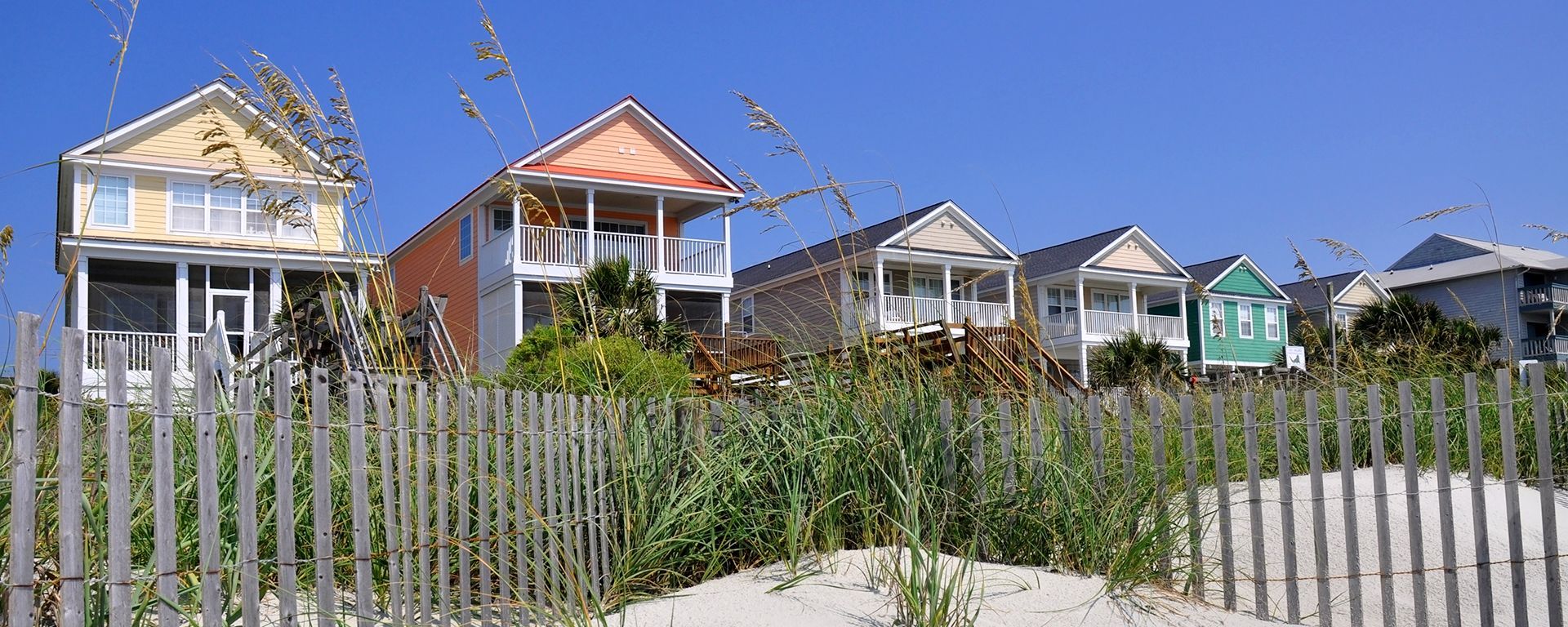 A row of colorful beach houses with porches behind a wooden sand fence and grassy dunes under a clear blue sky.