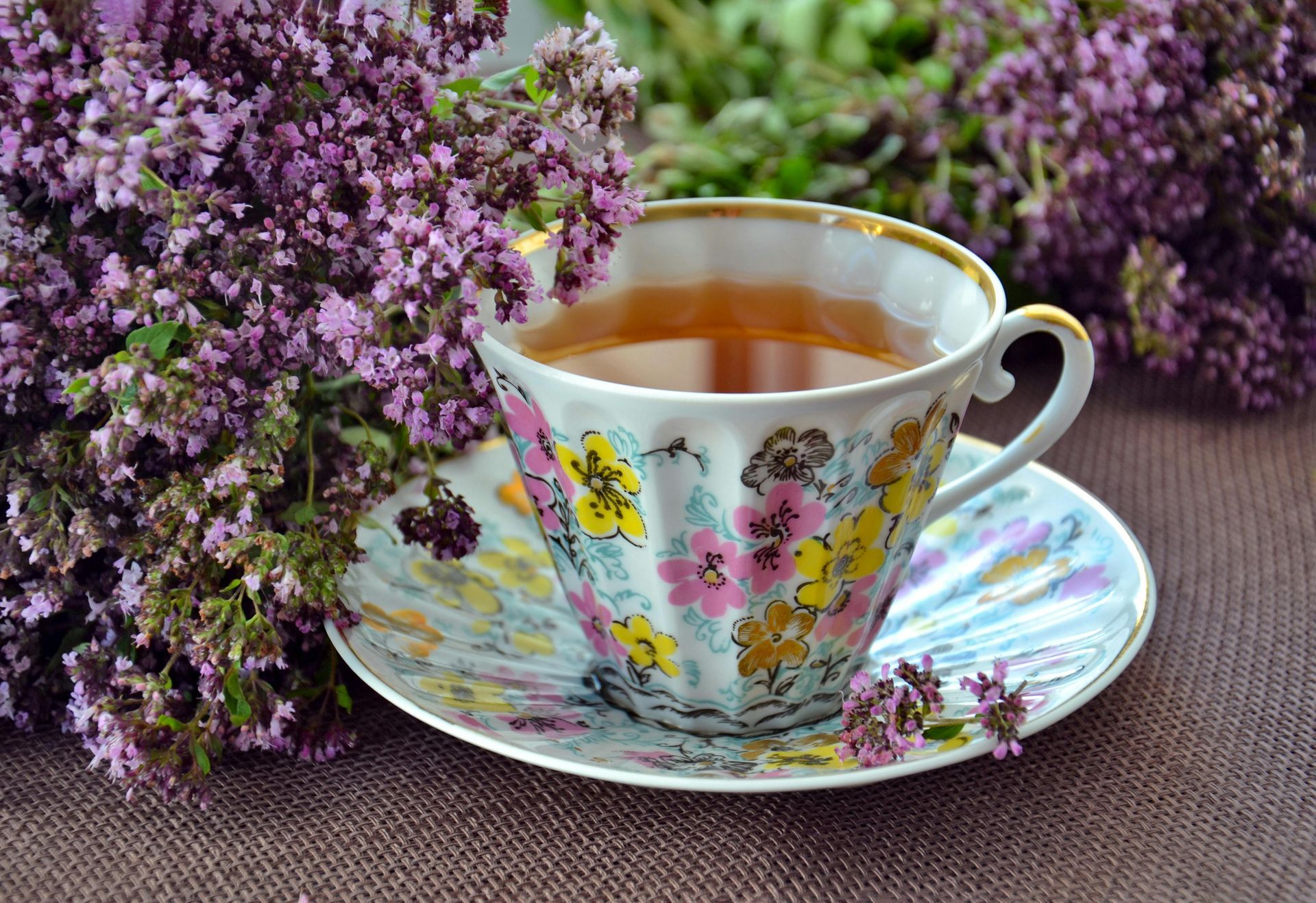 A cup of tea is on a saucer next to purple flowers.