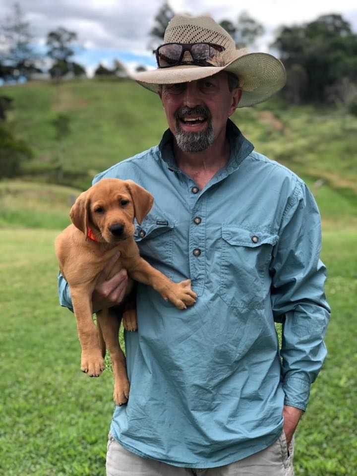 Man in hat holding a golden puppy in a grassy field.