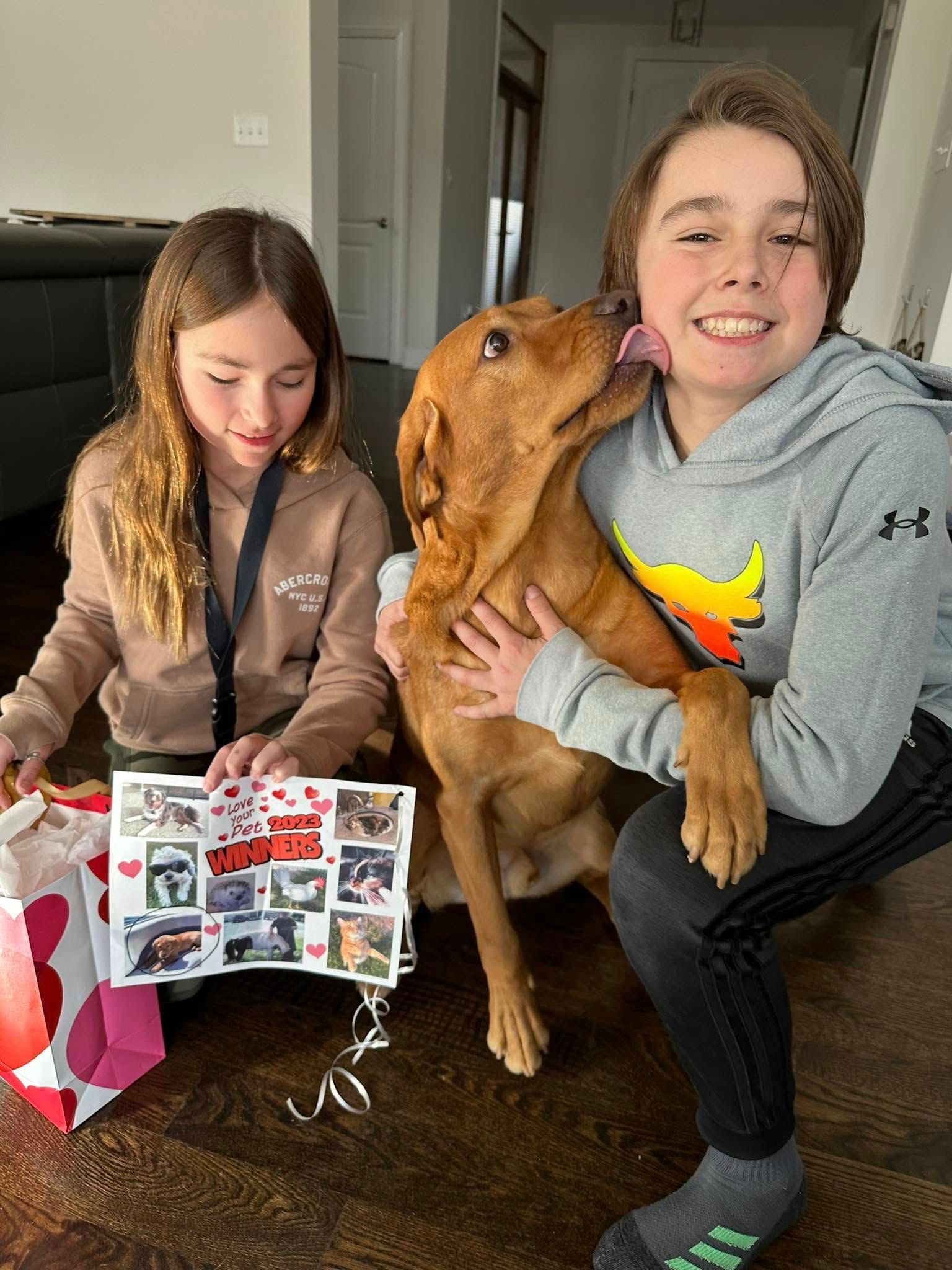 Girl and boy with a dog; the dog licks the boy's face. The girl holds a photo album. Indoors, natural light.