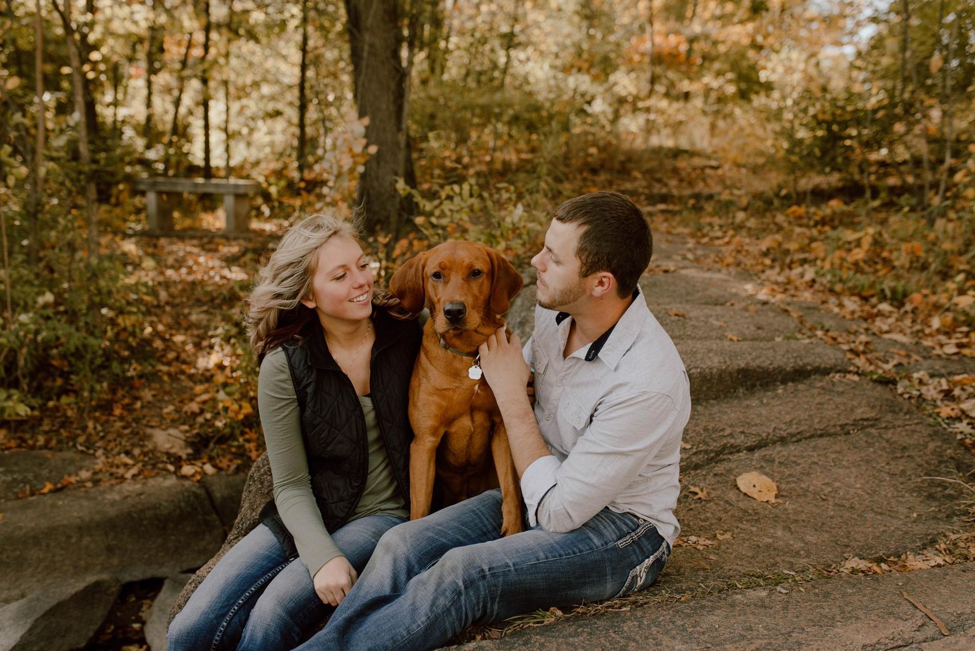 Couple with dog sitting on rock in autumn park; woman in vest, man petting dog.