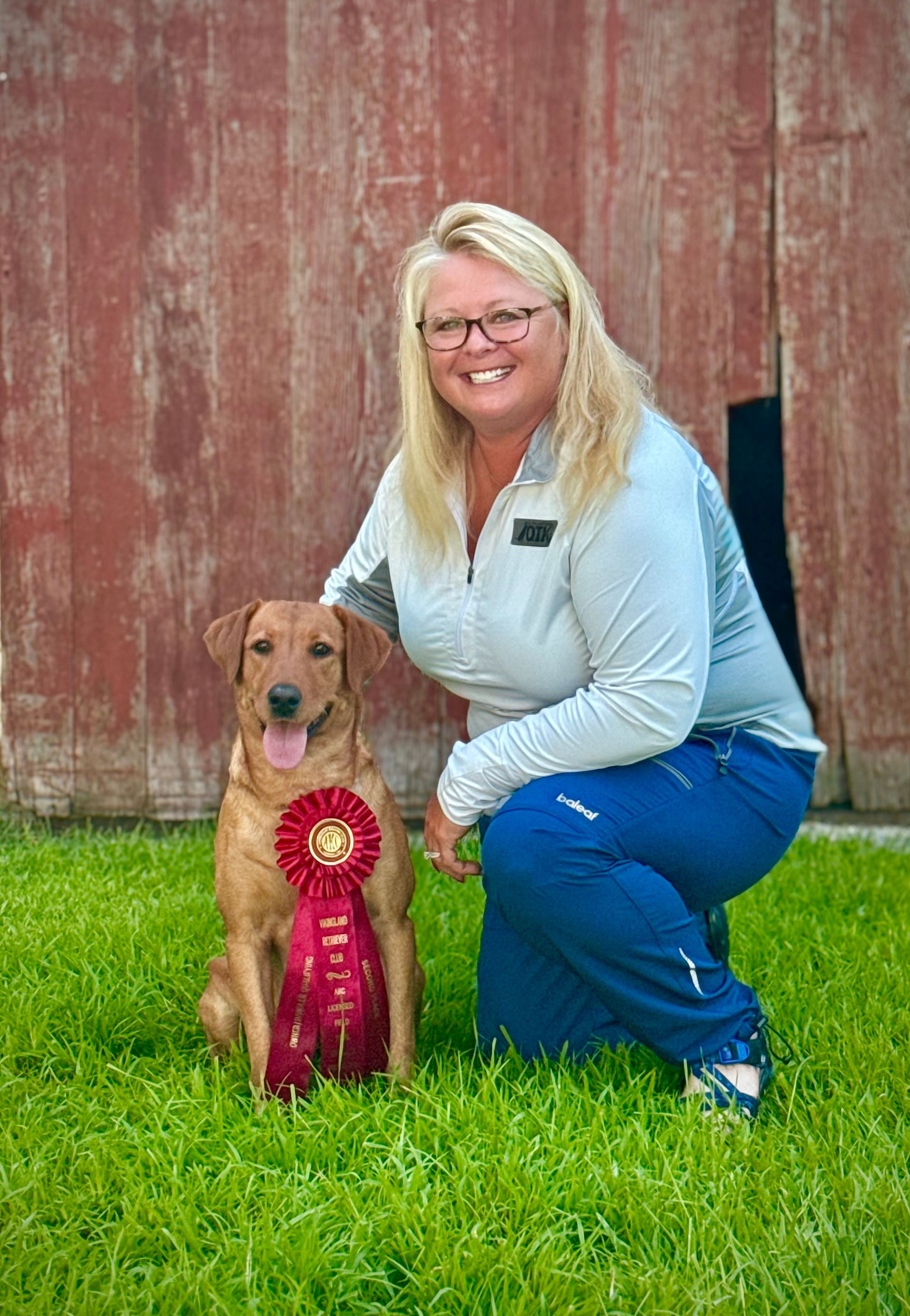 Woman kneels beside a reddish-brown dog with a ribbon. They are on grass, with a red barn in the background.