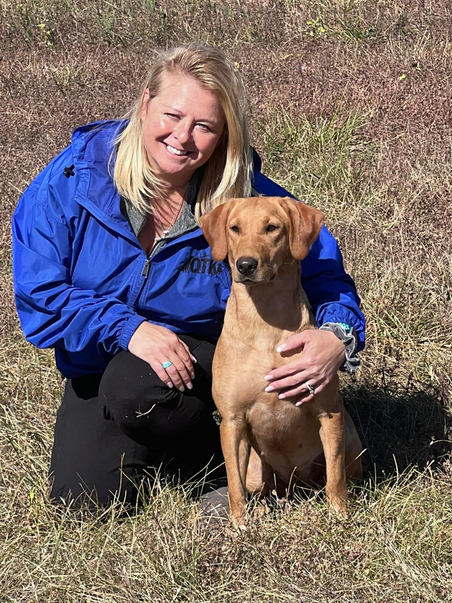 Woman kneels, smiles, and pets a brown dog outdoors on dry grass, both looking at the camera.