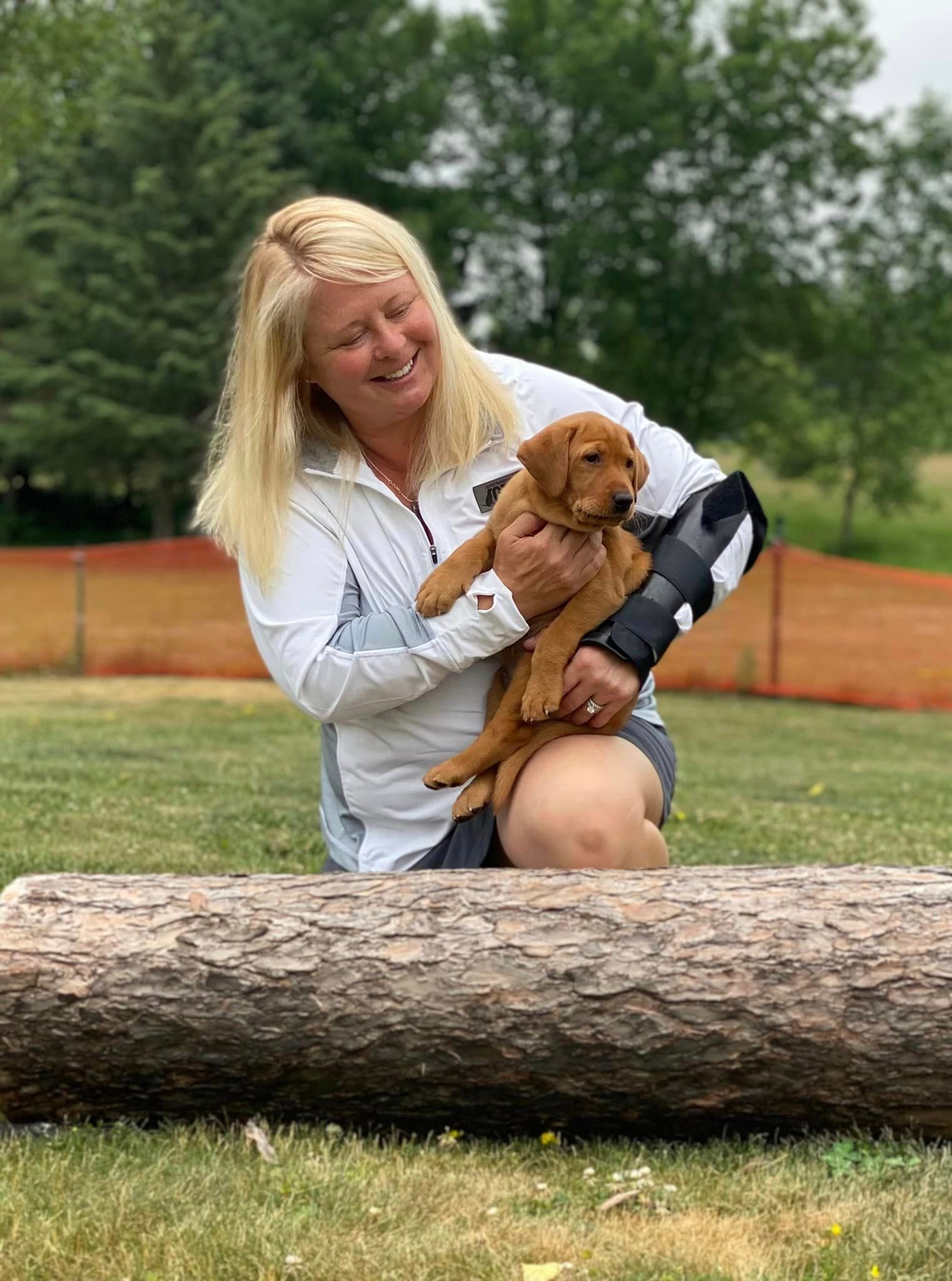 Woman with long blonde hair smiles while holding a small, brown puppy outdoors. A large log is in front of them.