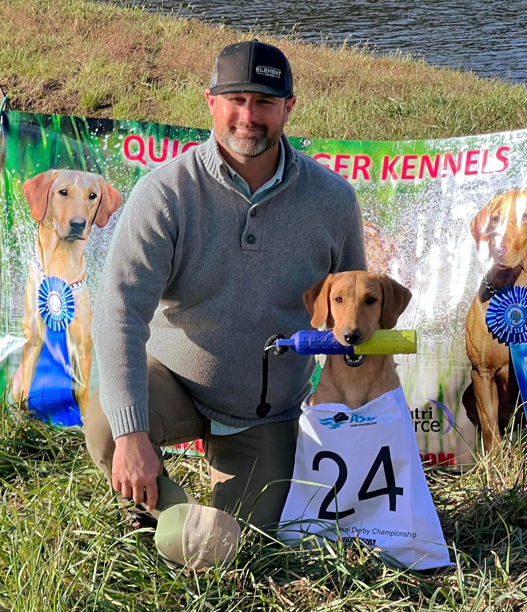 Man kneels beside a golden retriever holding a toy, wearing a bib with number 24.