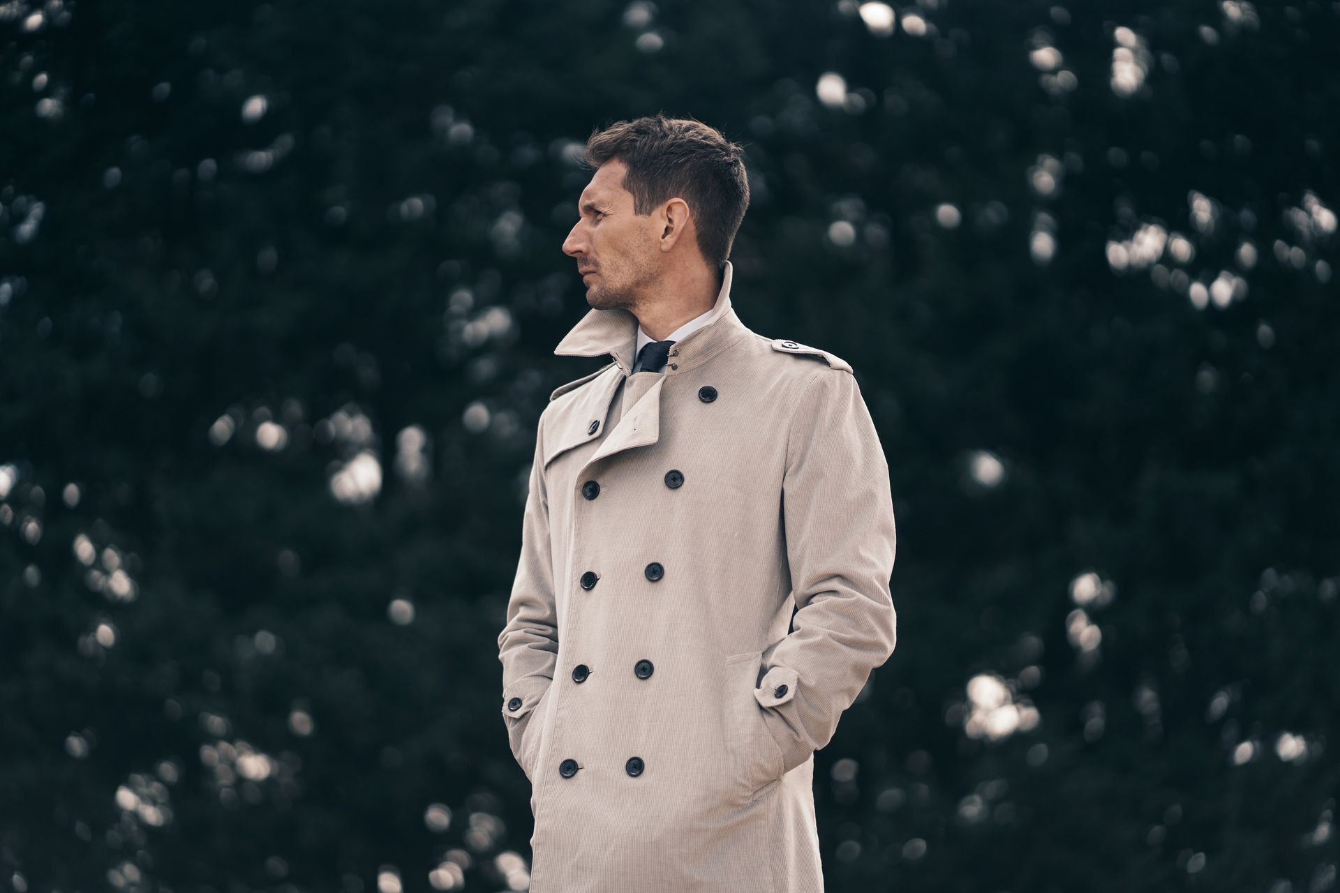 A man in a trench coat is standing in front of trees.