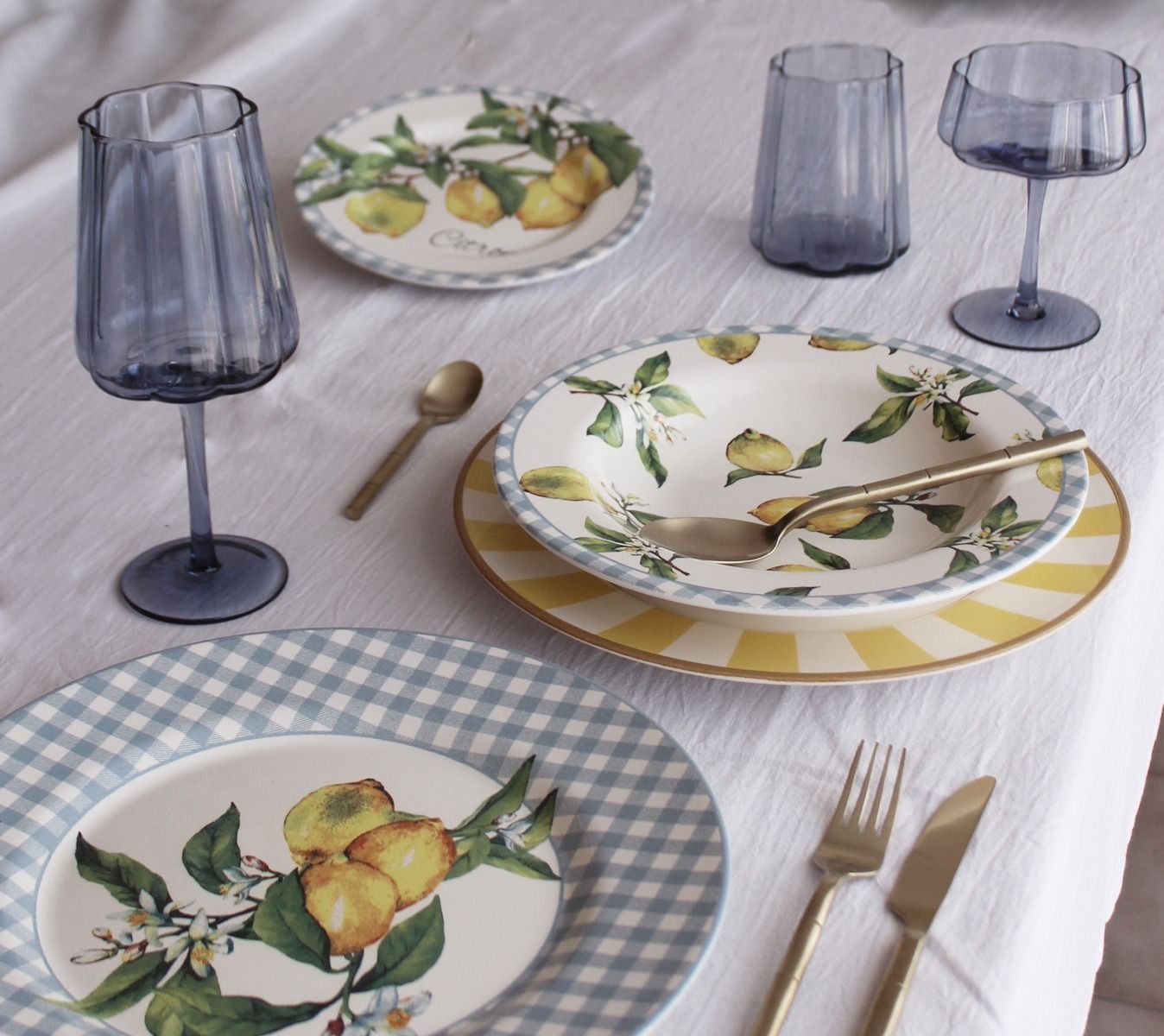 Mesa con vasos azules, platos con temática de limón, cubiertos dorados y mantel blanco.