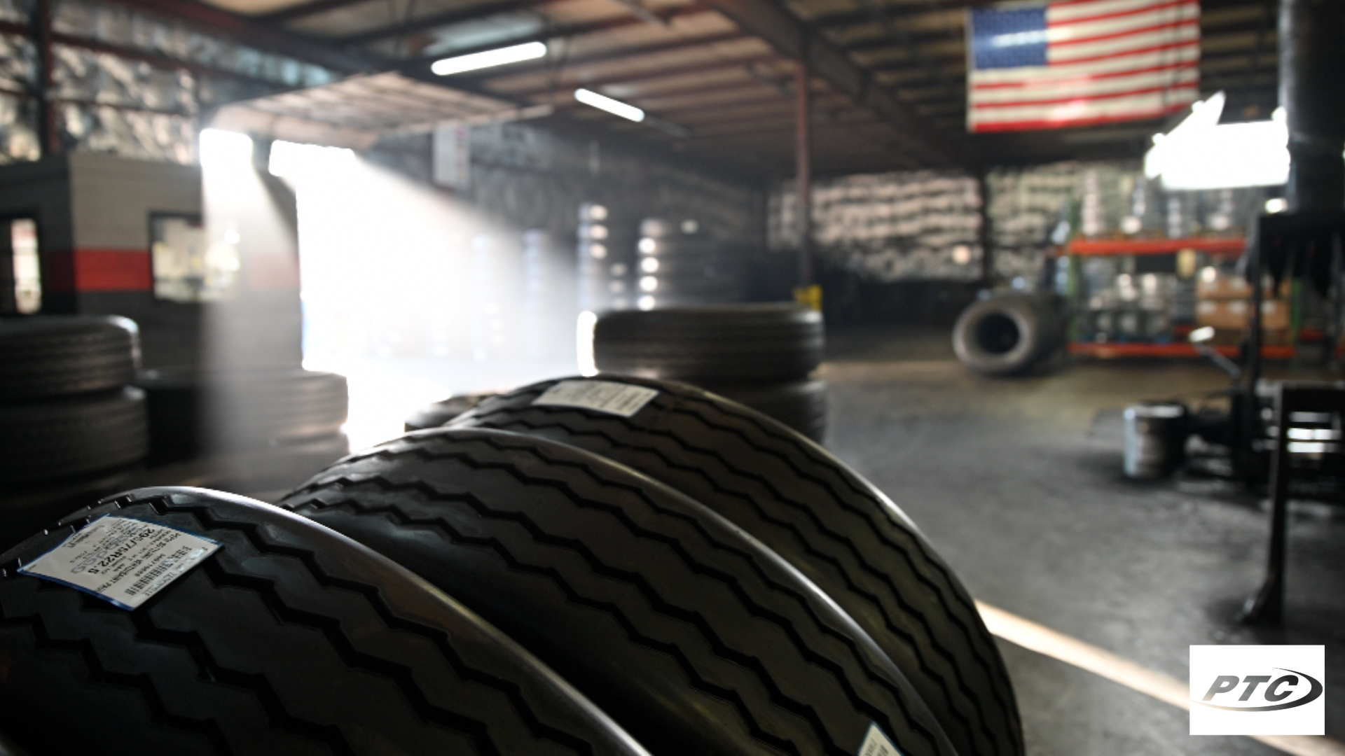 Tires stacked in a garage with sunlight streaming through, American flag in the background.