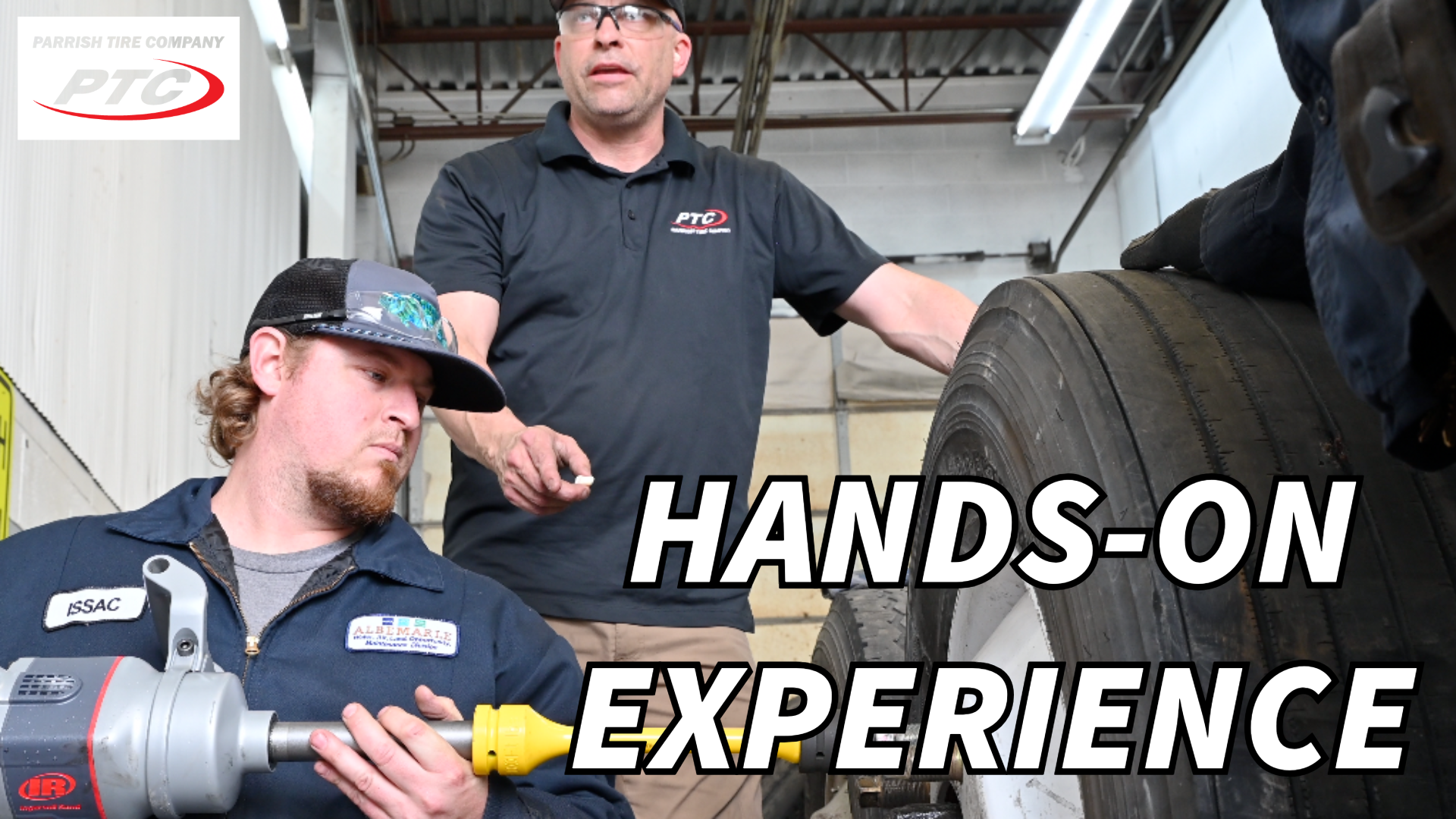 Two mechanics working on a truck tire. One uses a tool, the other supervises.