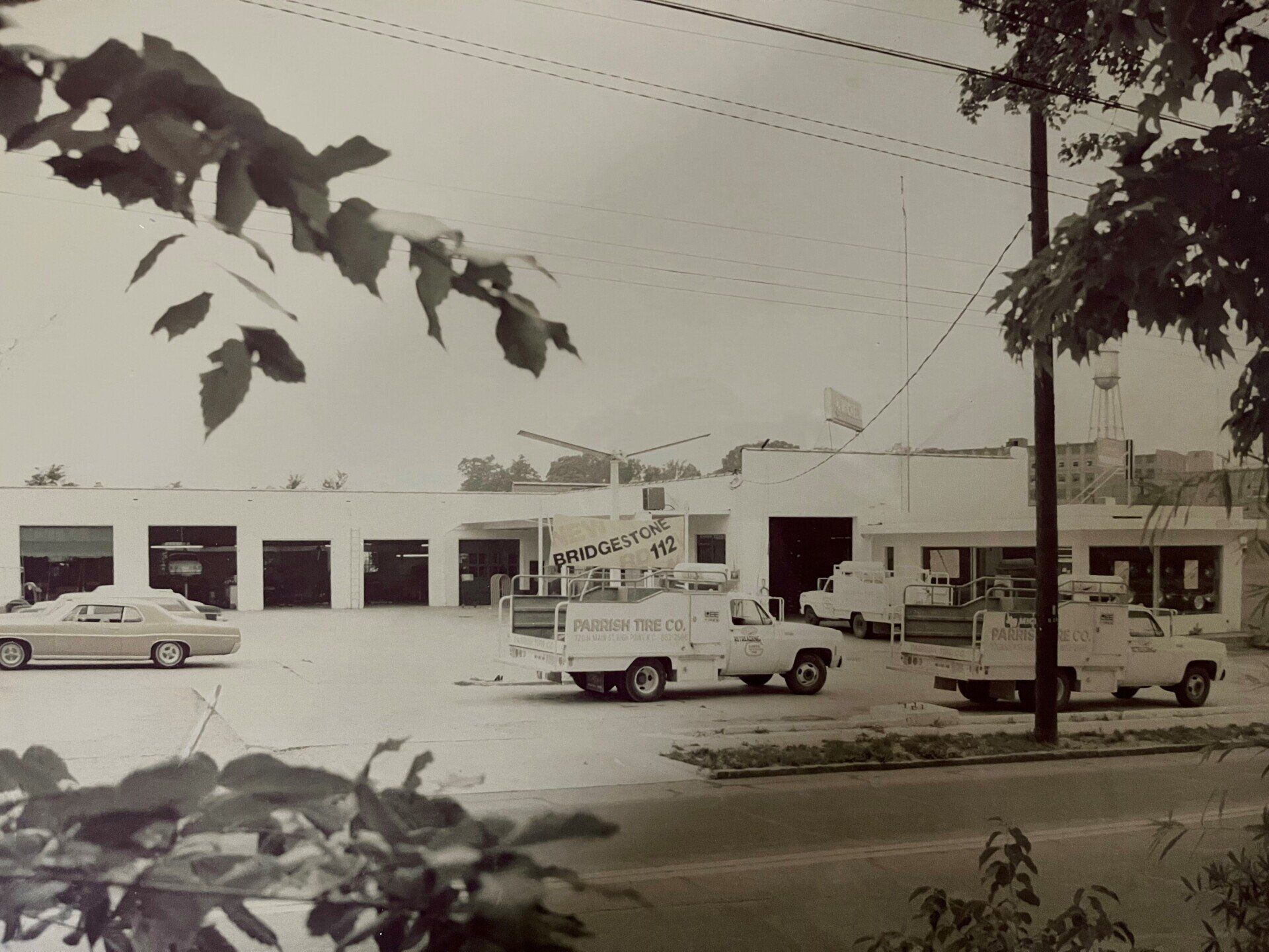 Black and white photo of a building with garage doors, service trucks parked in front, and a car.