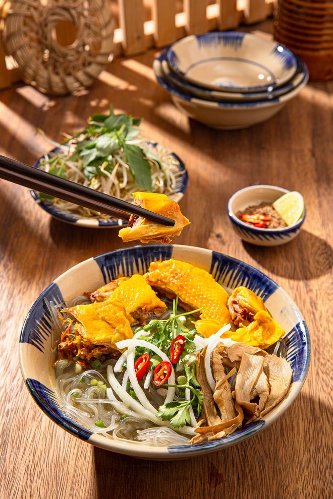 A bowl of Vietnamese chicken noodle soup with fresh herbs, onions, and a side of chili sauce on a wooden table.