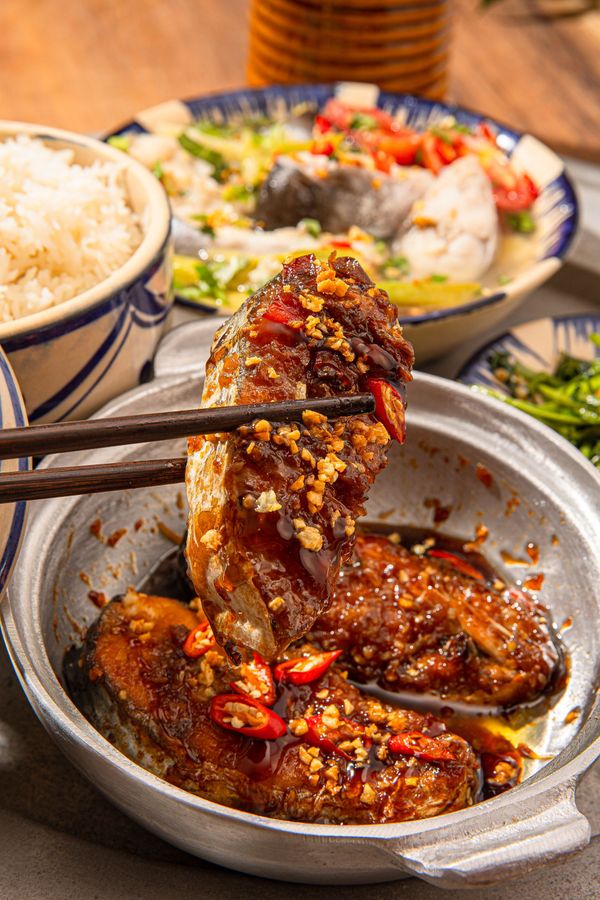 Chopsticks lift a piece of glazed, chili-topped fish from a metal pot, accompanied by a bowl of rice and a vegetable dish.