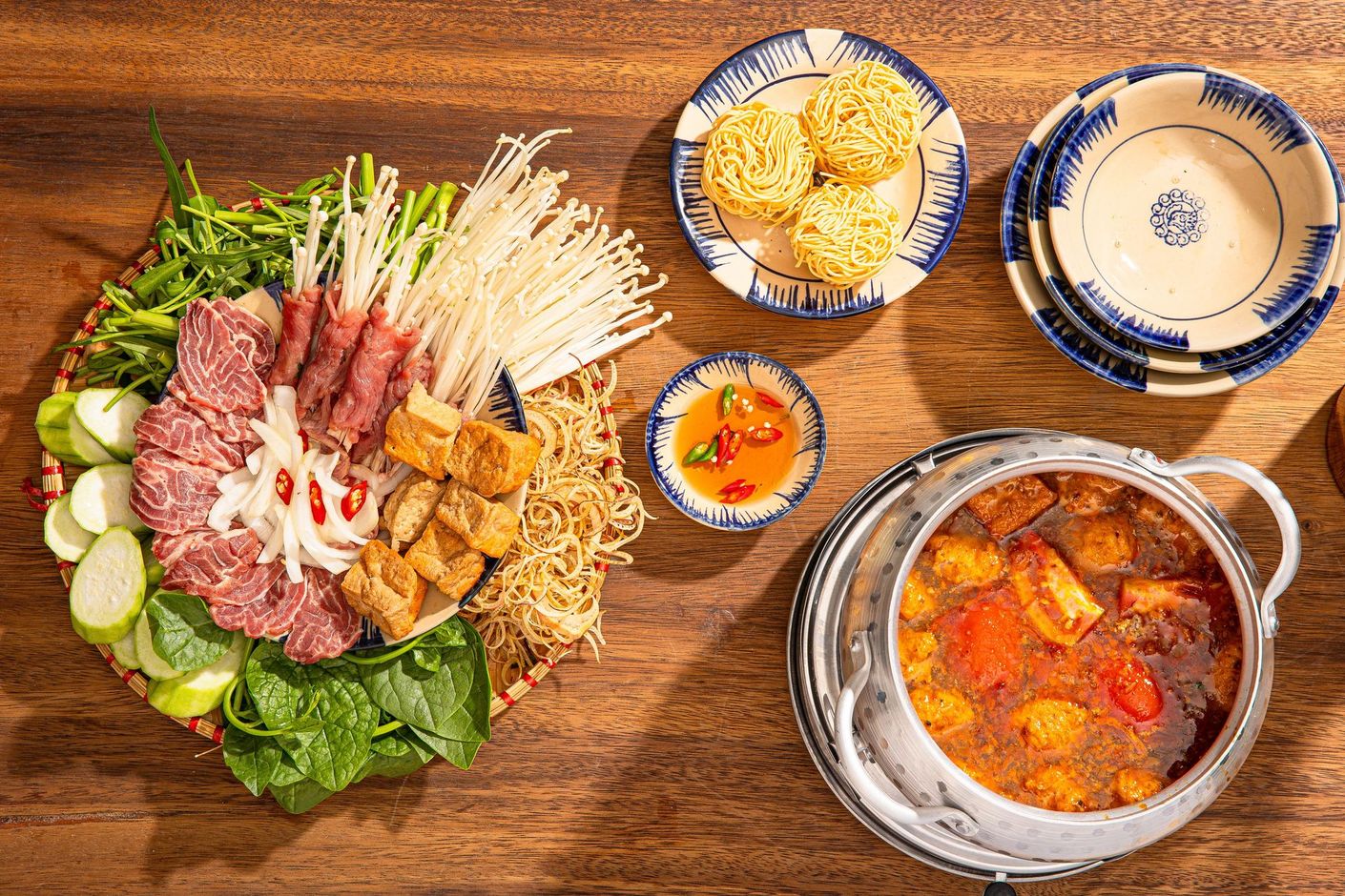 A top-down view of a Vietnamese hot pot meal with raw beef, tofu, mushrooms, greens, noodles, and a pot of broth.
