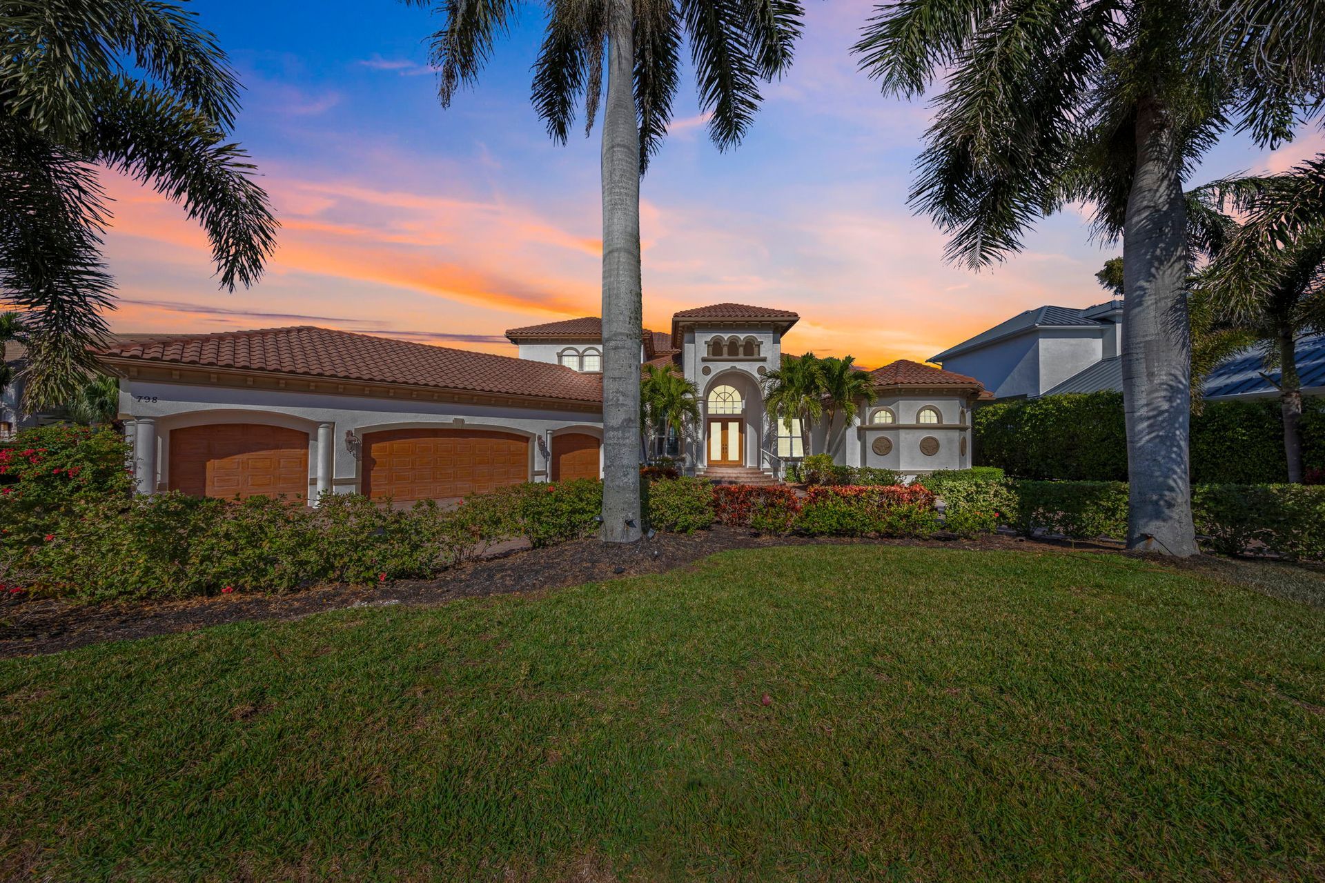 Suburban home at dusk with red tile roof, palm trees, and a manicured front lawn.