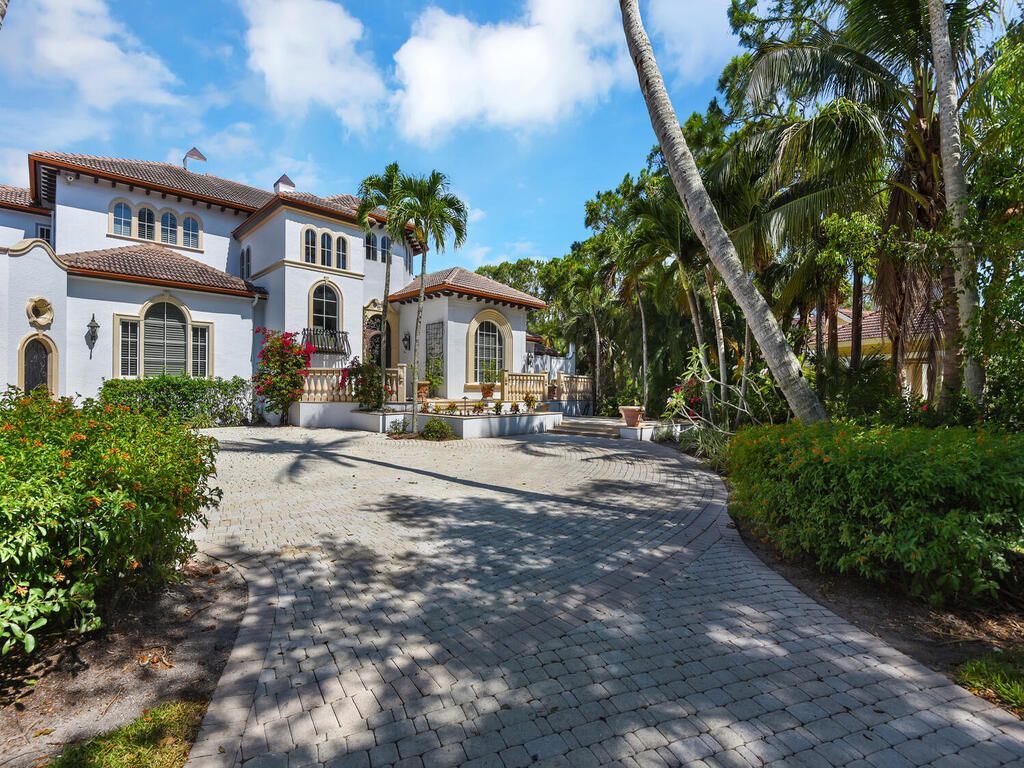 Spanish-style villa with white stucco, red tile roof, and a brick driveway framed by palm trees