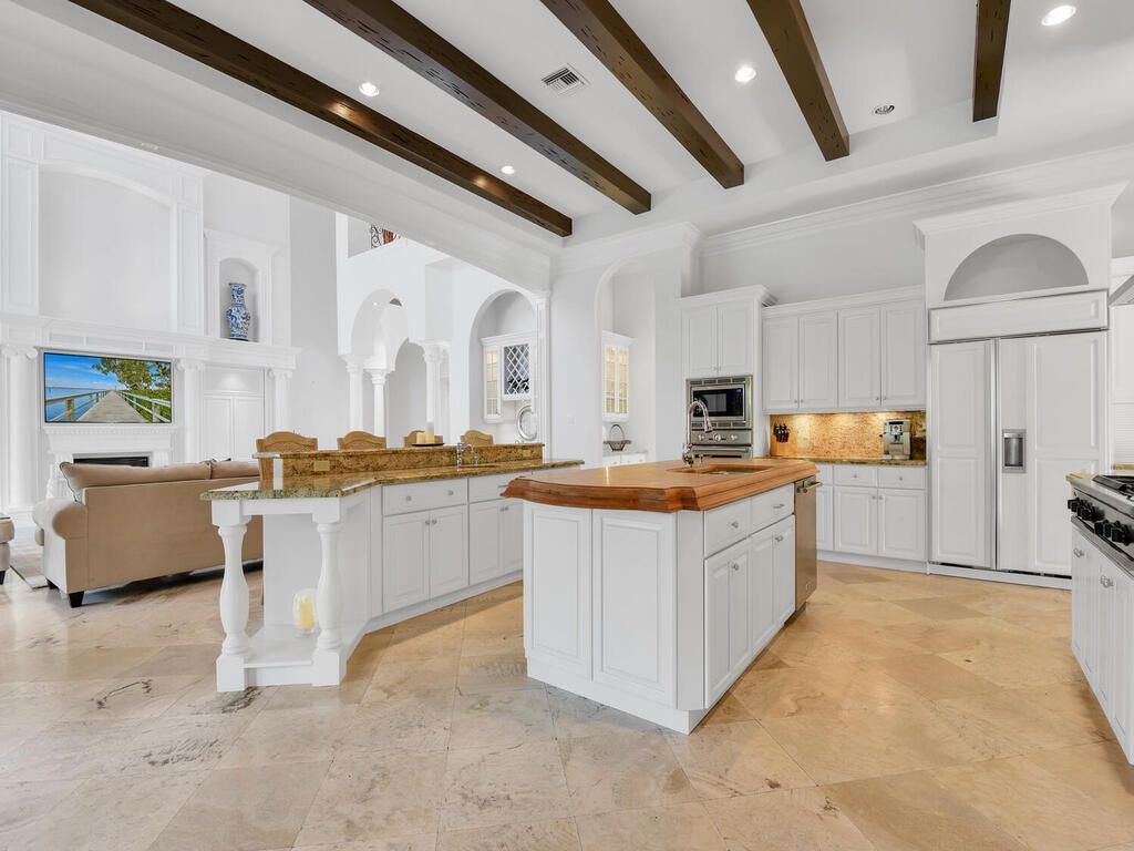 Bright white luxury kitchen with wood beam ceiling, island, arched cabinets, and beige stone floor