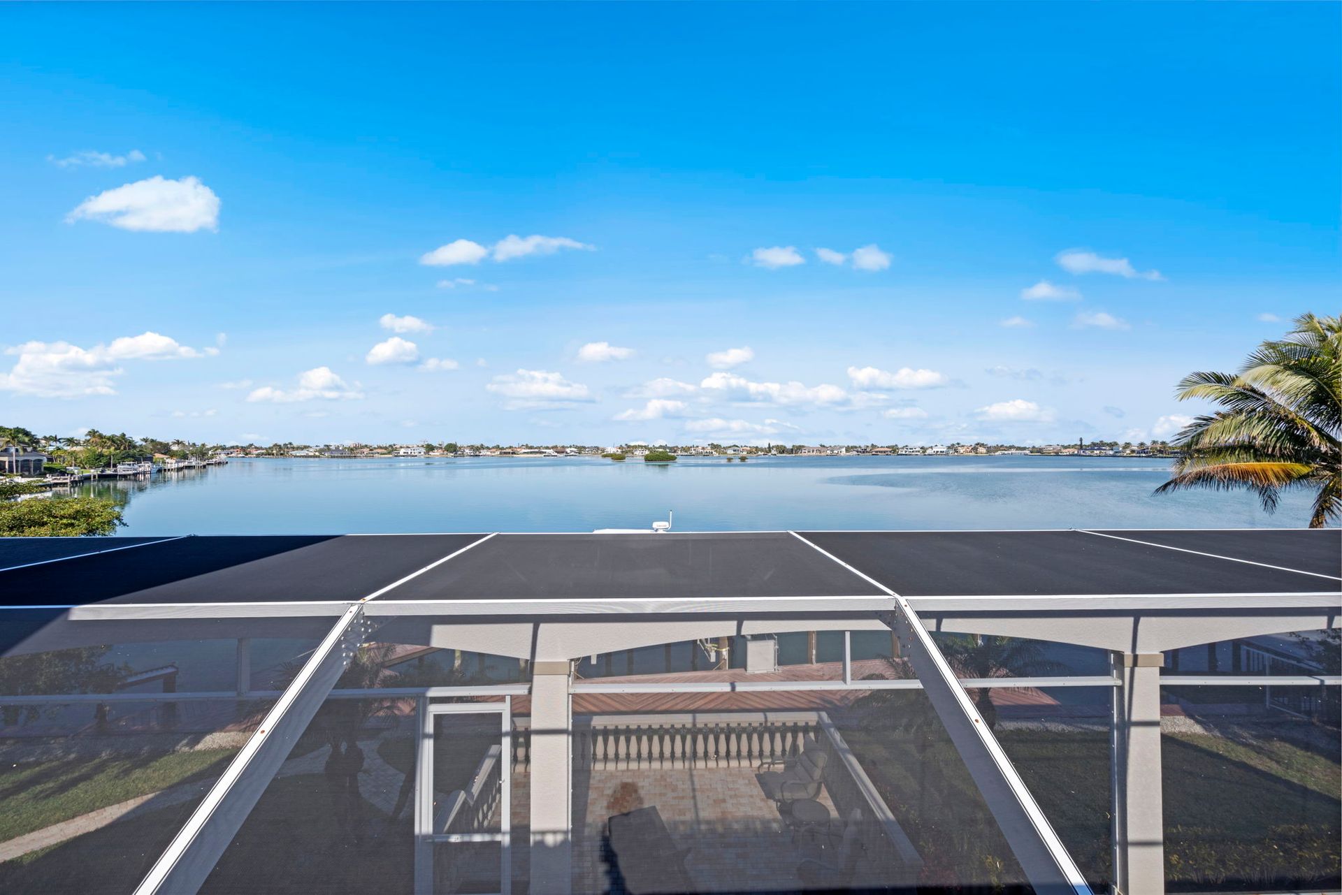 Sunny waterfront view from a balcony with a glass railing and calm water under a blue sky.