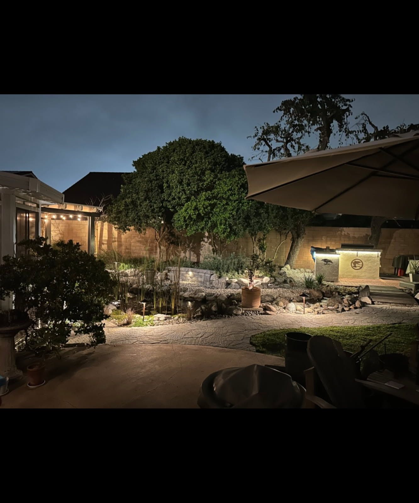 Nighttime patio garden in Corona, CA, with lit pathways, large tree, stone landscaping, and a shaded seating area